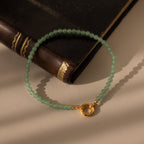 The Beaded Jade Clasp Bracelet, featuring green jade beads and a gold clasp, rests atop a brown leather book bathed in soft sunlight.