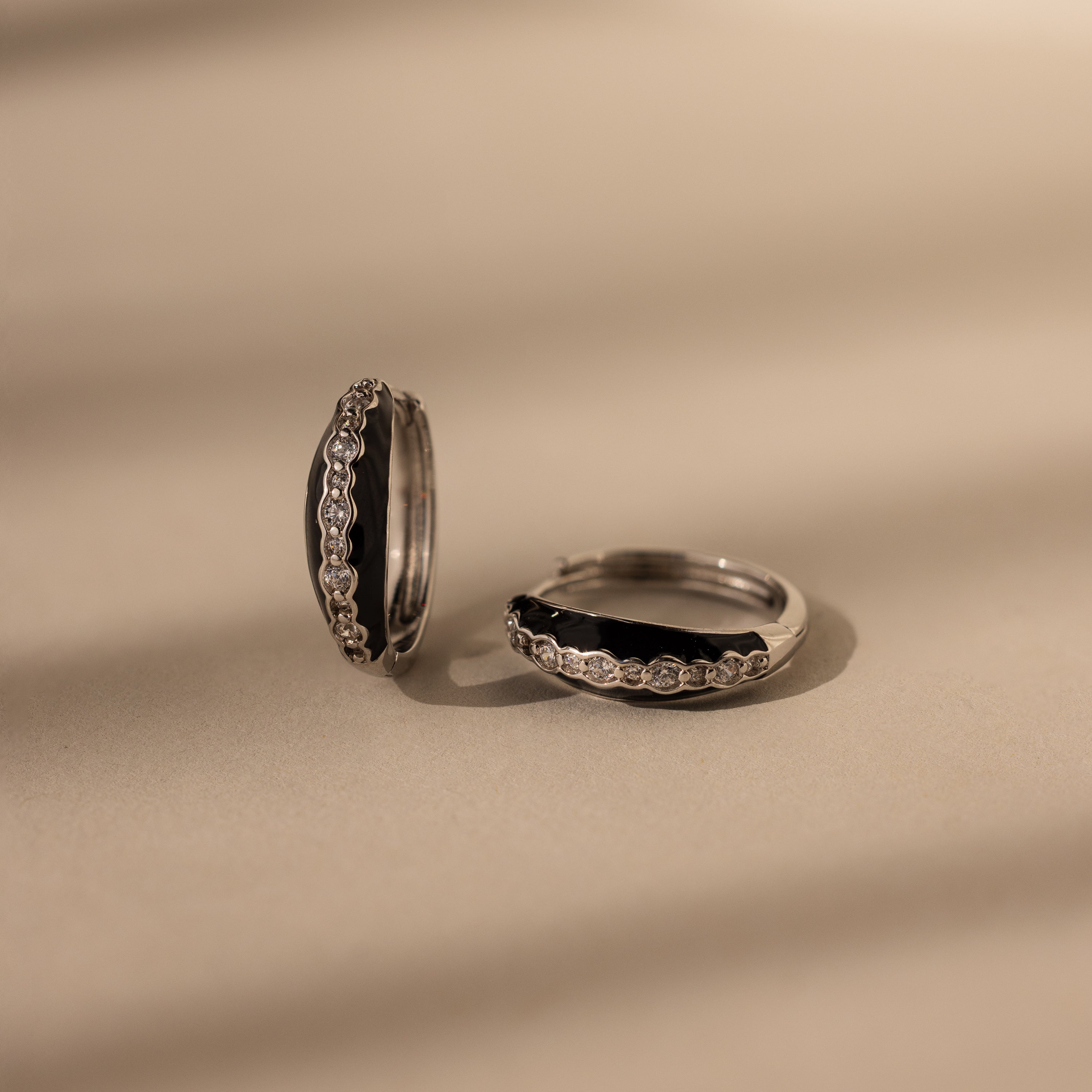 The Noir Pave Dome Hoops in Silver are statement earrings featuring black enamel and small clear stones, shown on a beige surface.