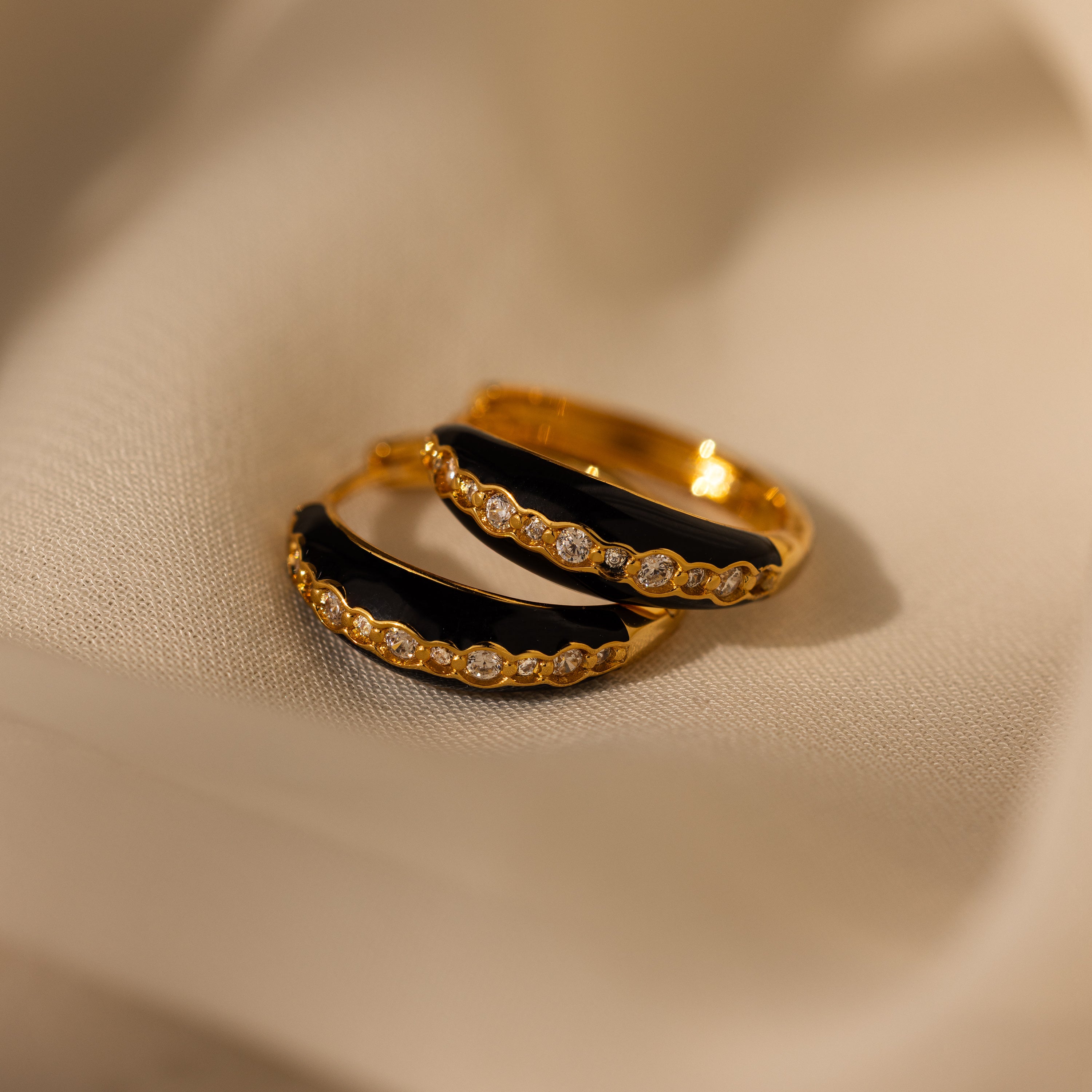 Noir Pave Dome Hoops in 18K Gold feature black enamel accents and small diamonds, displayed on soft beige fabric—elegant statement earrings for any occasion.