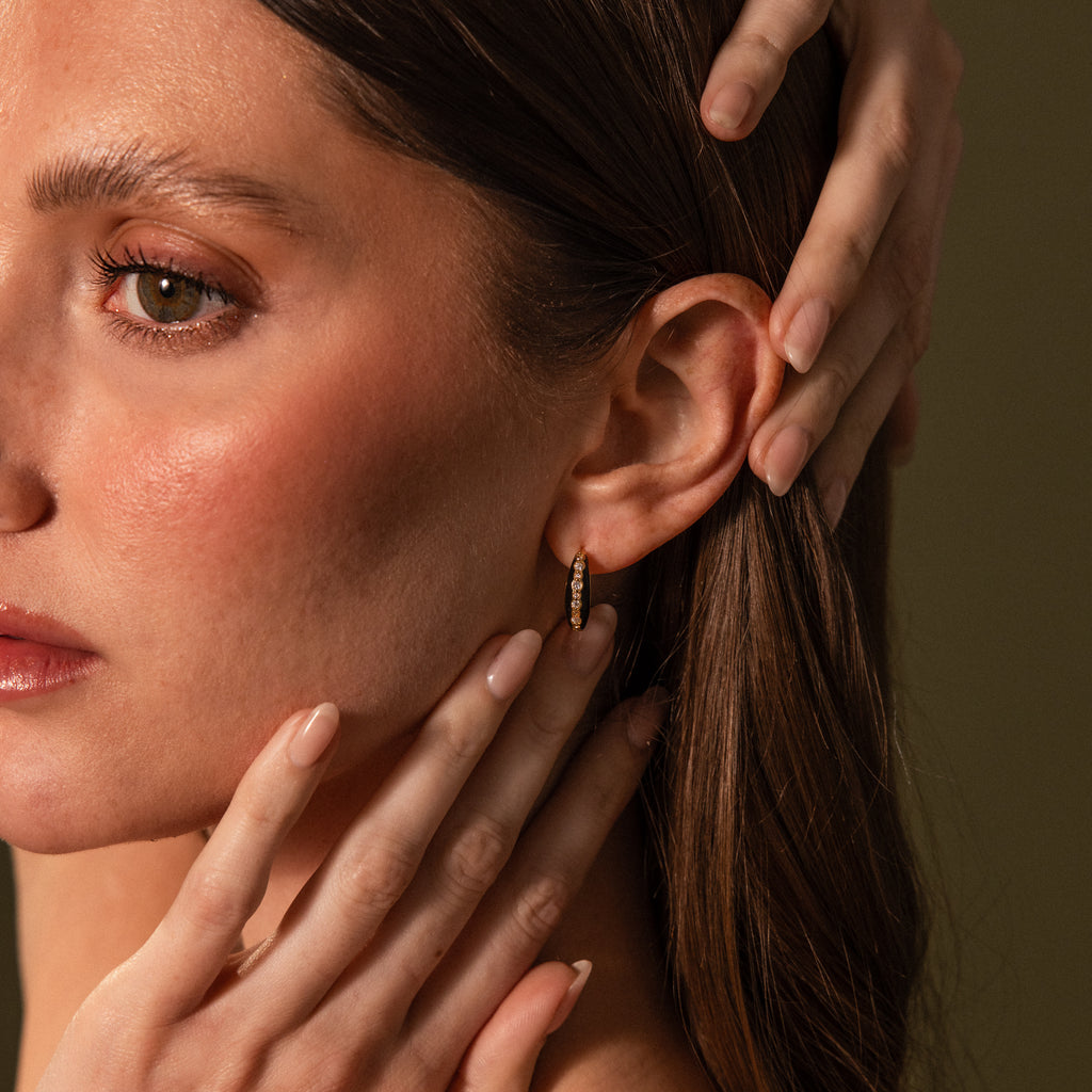 A woman with brown hair touches her face, highlighting the Noir Pave Dome Hoops in 18K Gold, statement earrings featuring a subtle enamel black accent, set against a plain background.