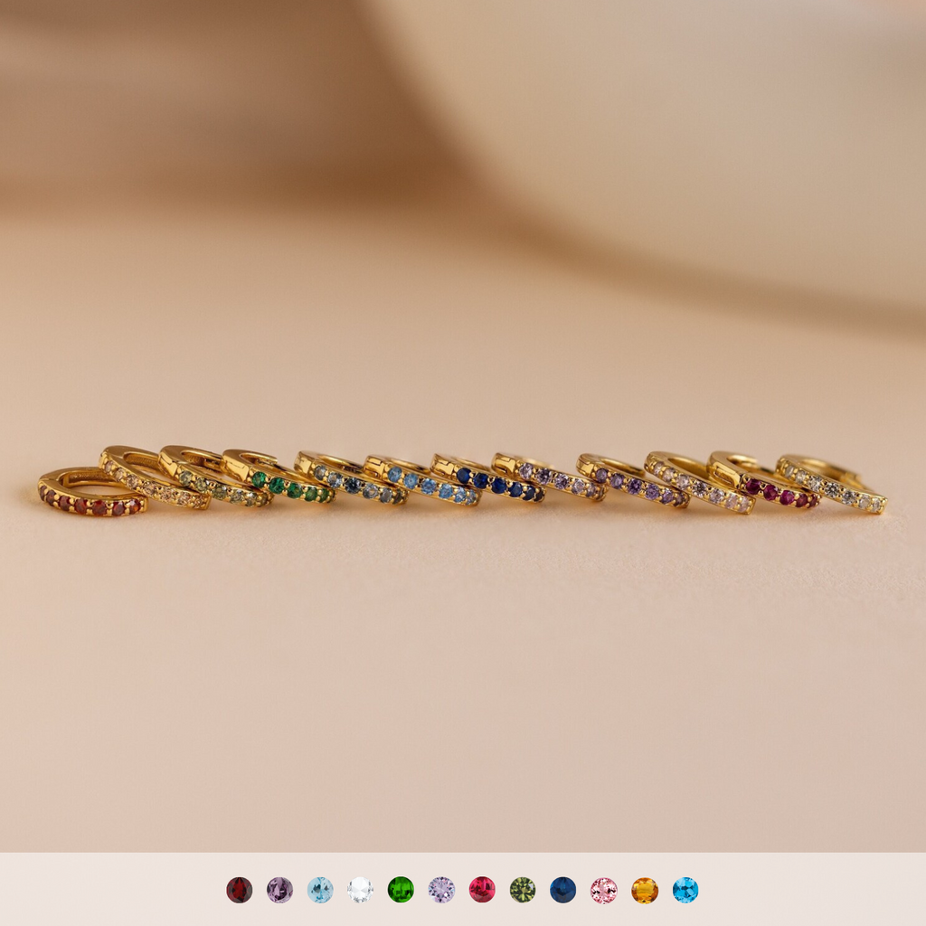 Ten gold rings with vivid gemstones are lined up on a beige surface, each featuring a unique color—pair them with Dainty Pave Birthstone Huggies for a bright, coordinated look.