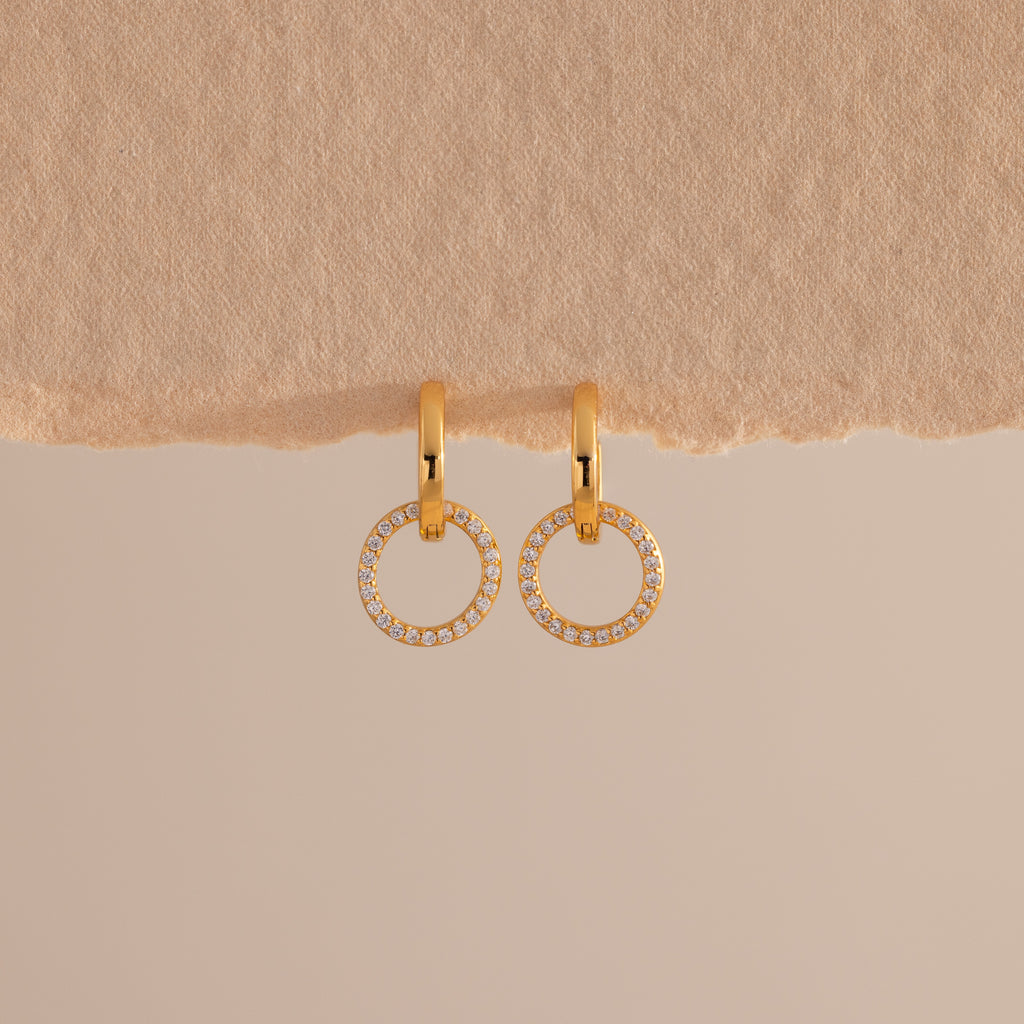 A pair of elegant gold huggie earrings, each featuring a small open circle drop encrusted with sparkling pave stones hang against a minimal, soft beige background with a textured paper edge at the top.