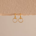 A pair of elegant gold huggie earrings, each featuring a small open circle drop encrusted with sparkling pave stones hang against a minimal, soft beige background with a textured paper edge at the top.