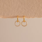 A pair of elegant gold huggie earrings, each featuring a small open circle drop encrusted with sparkling pave stones hang against a minimal, soft beige background with a textured paper edge at the top.
