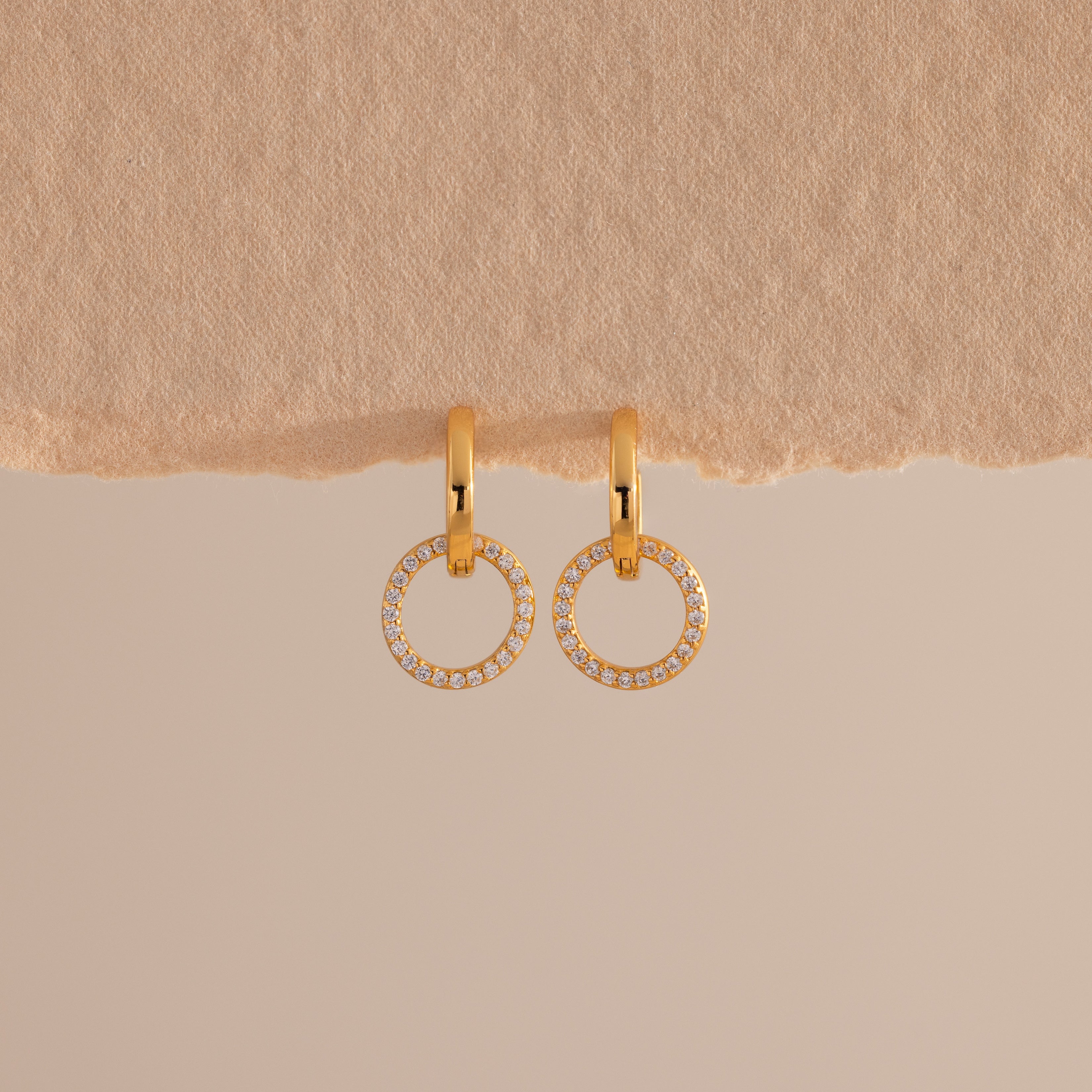 A pair of elegant gold huggie earrings, each featuring a small open circle drop encrusted with sparkling pave stones hang against a minimal, soft beige background with a textured paper edge at the top.