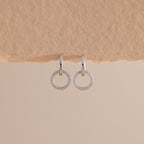 A pair of elegant silver huggie earrings, each featuring a small open circle drop encrusted with sparkling pave stones hang against a minimal, soft beige background with a textured paper edge at the top.