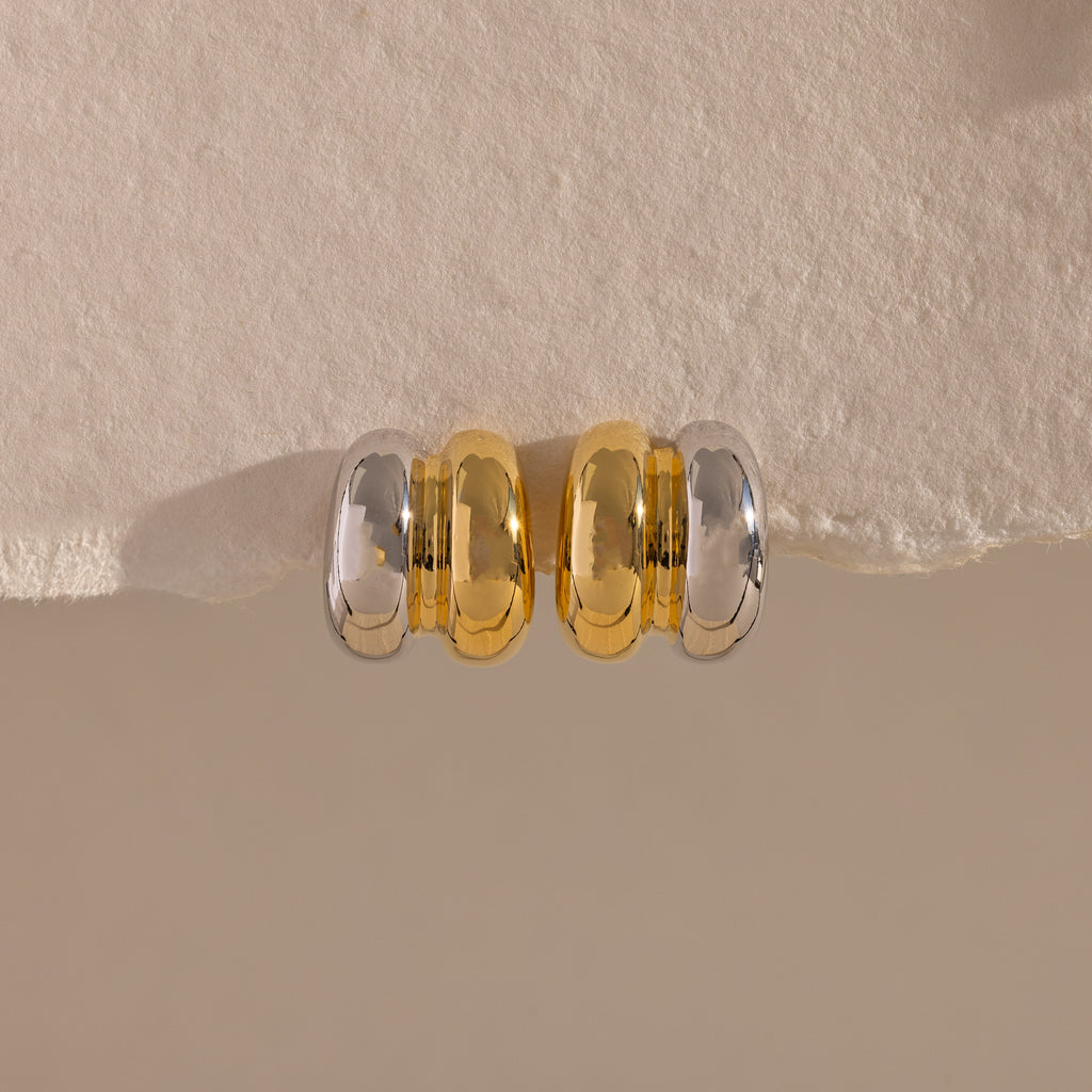 Mixed Metal Puff Studs—one silver, one gold—rest side by side on a textured beige surface. Perfect for fans of modern earrings and minimal jewelry.