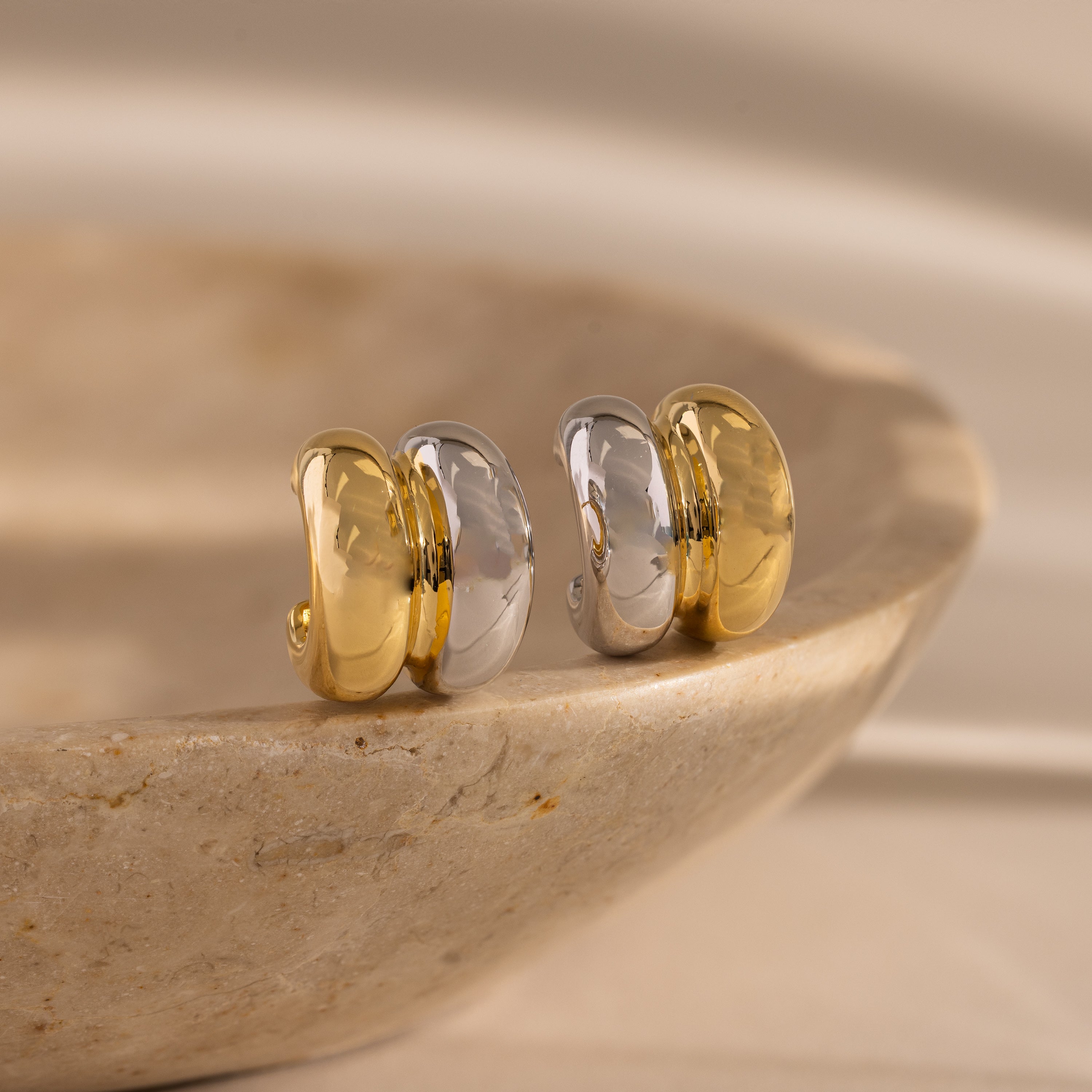 The Mixed Metal Puff Studs, featuring bold gold and silver tones, sit atop a smooth stone surface—perfect for fans of minimal jewelry.