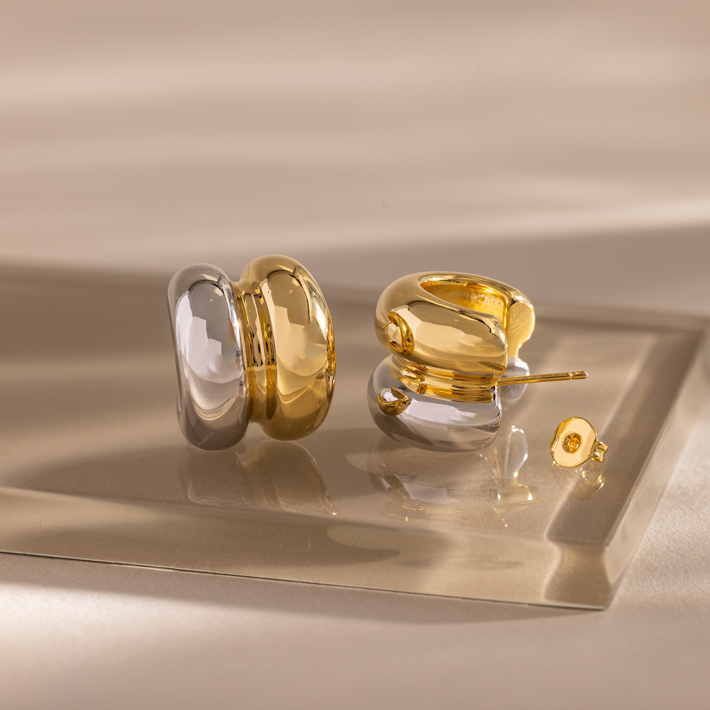 The Mixed Metal Puff Studs—one silver, one gold—are shown on a beige reflective surface under soft lighting, highlighting their modern, minimal jewelry style.