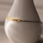 The Mixed Metal Knot Bracelet, crafted from gold and silver snake chains, is elegantly displayed on a smooth white ceramic vase under soft lighting, capturing the essence of modern minimalism.