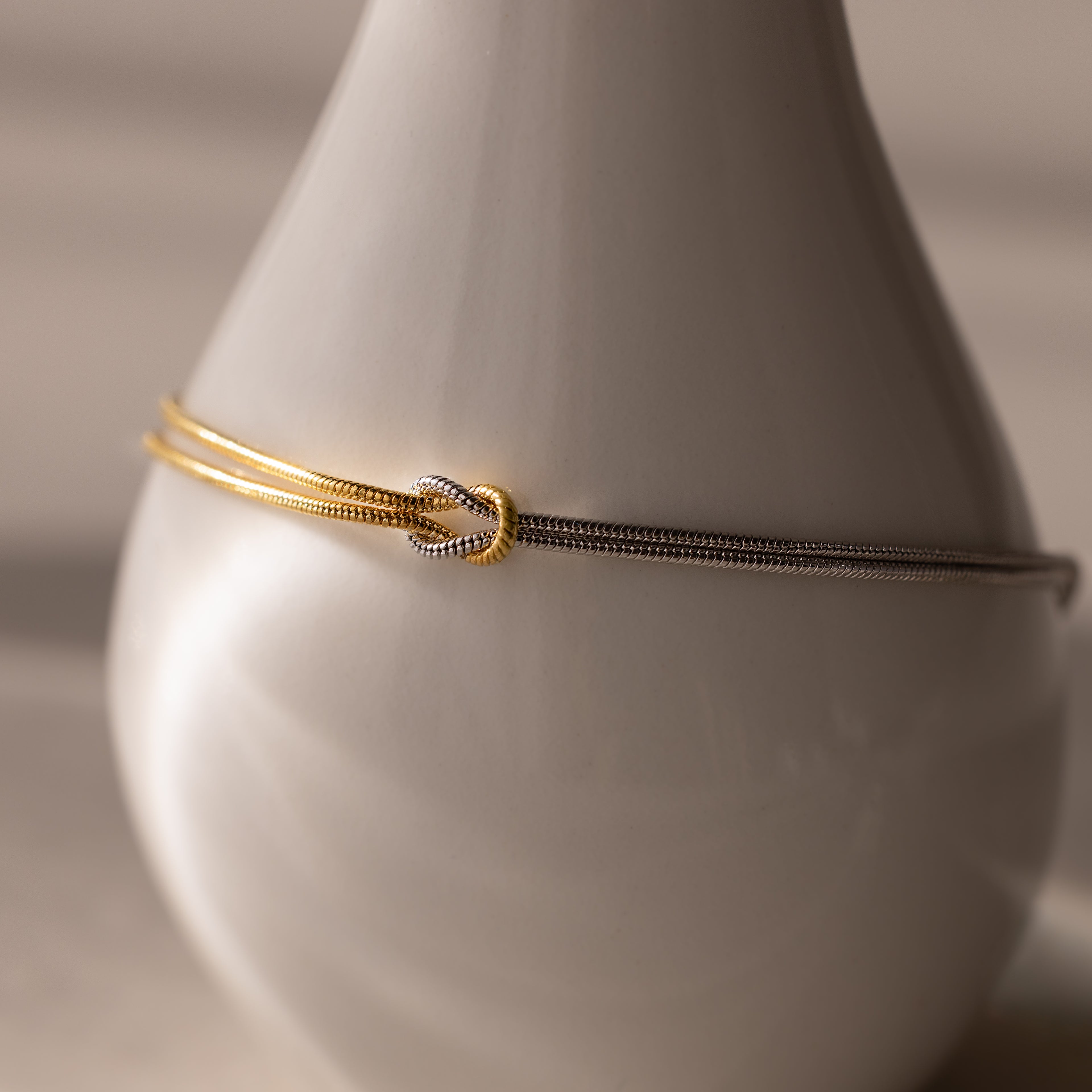 The Mixed Metal Knot Bracelet, crafted from gold and silver snake chains, is elegantly displayed on a smooth white ceramic vase under soft lighting, capturing the essence of modern minimalism.
