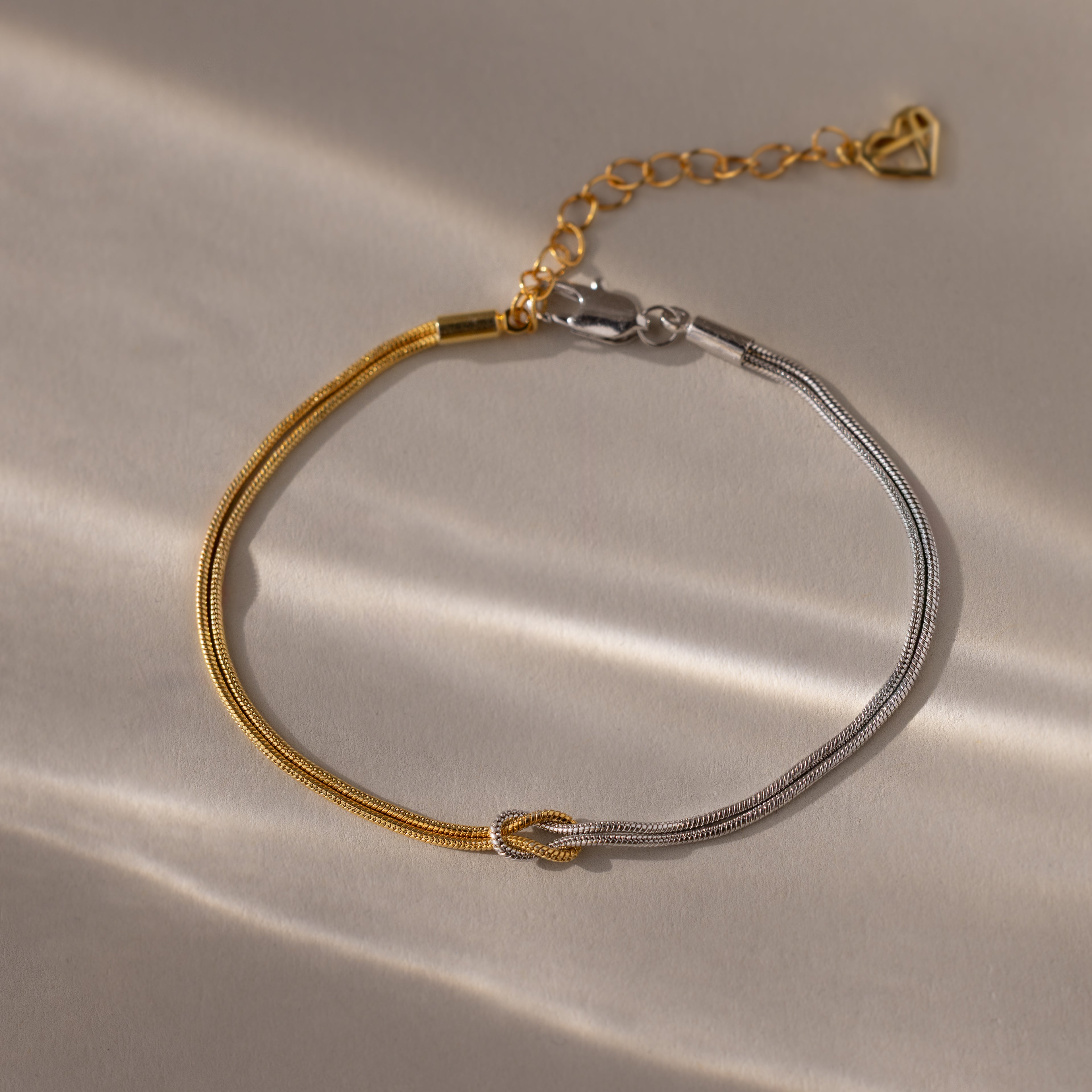 The Mixed Metal Knot Bracelet features two-tone gold and silver snake chains, a knot detail, and a heart charm—ideal for modern minimalists.