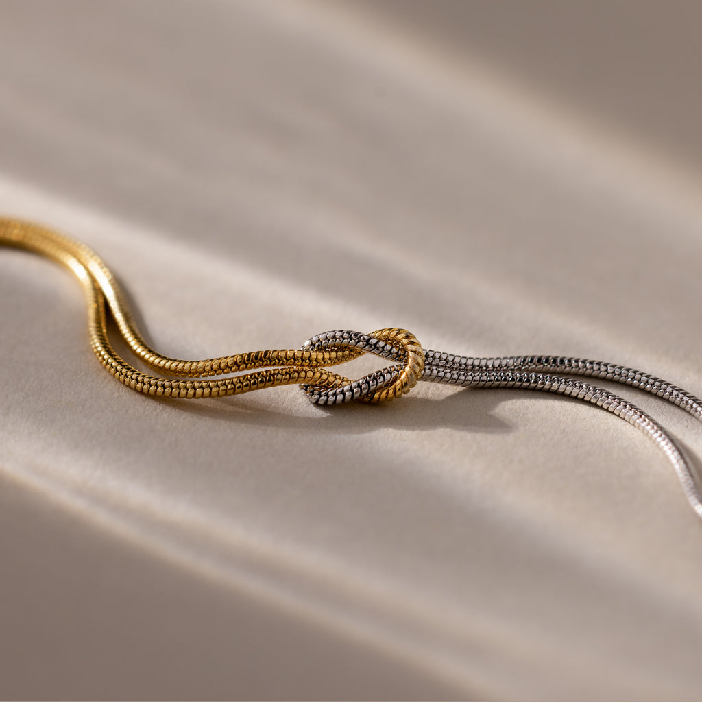 The Mixed Metal Knot Bracelet features intertwined gold and silver snake chains, inspired by modern minimalism, artfully knotted together for a striking mixed-metal look.