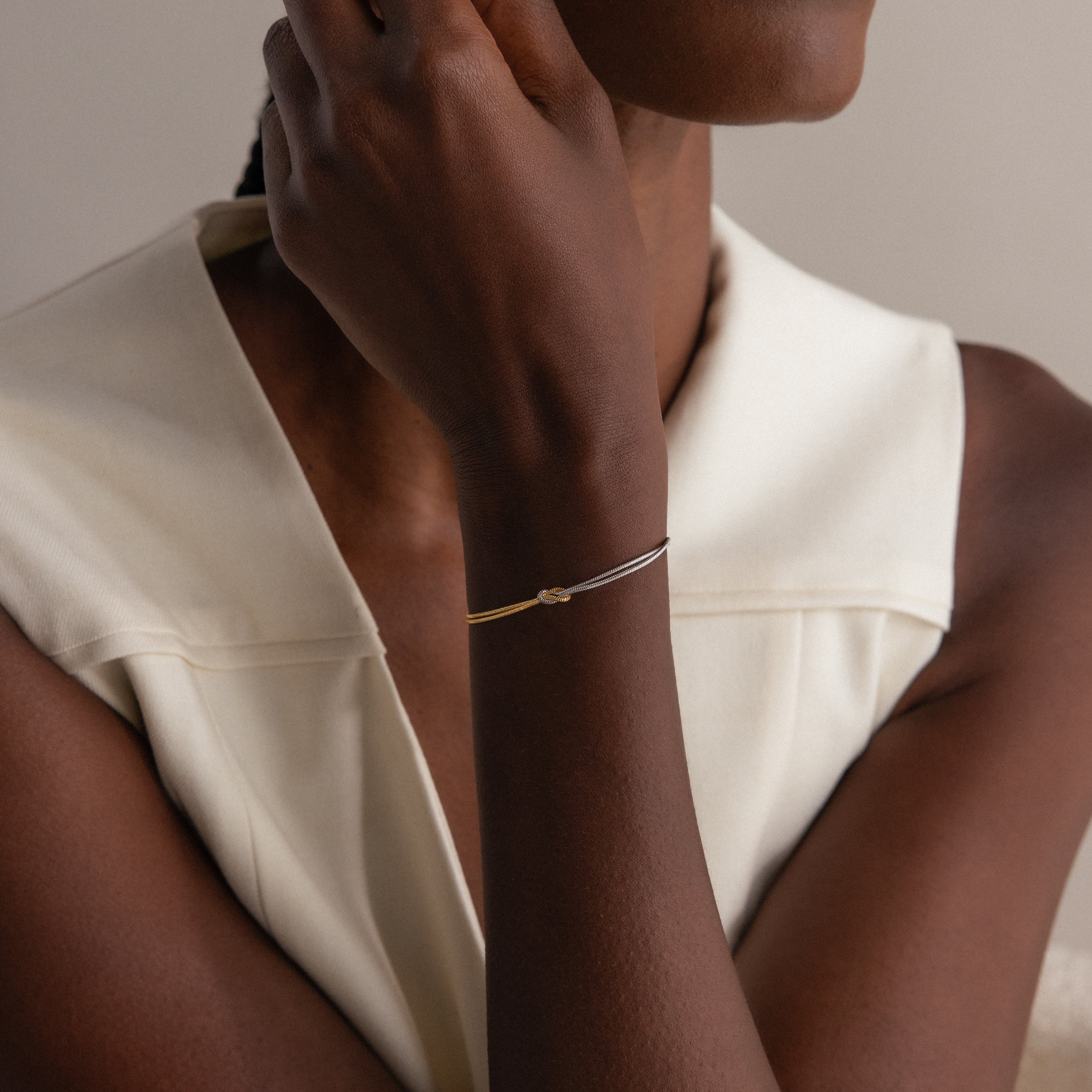 A woman wears a sleeveless white top and a Mixed Metal Knot Bracelet on her wrist, styled with gold and silver snake chains for a modern minimalist look.