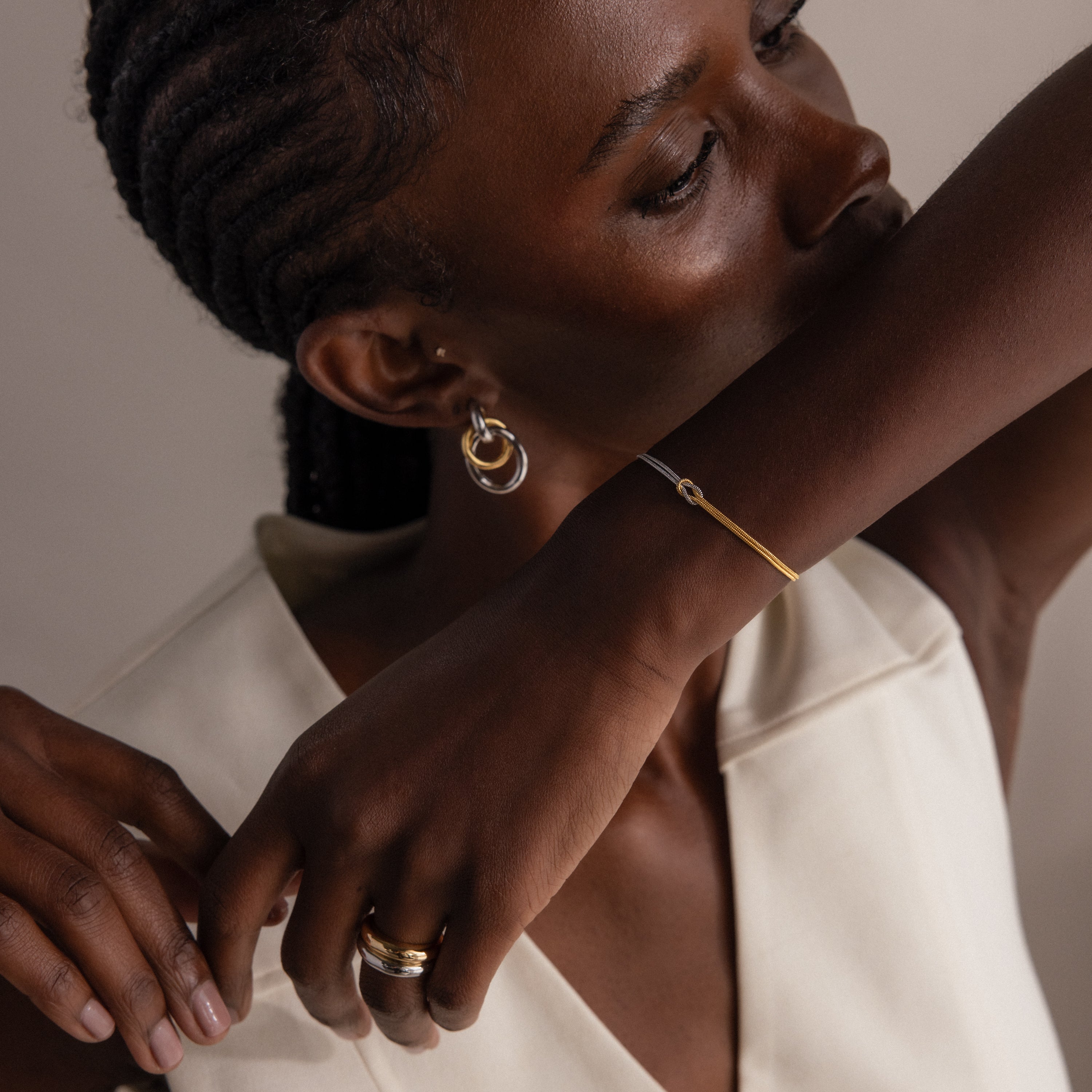 Woman with braided hair wearing gold jewelry and the Mixed Metal Knot Bracelet, paired with a sleeveless cream top, posing with her arm near her face.