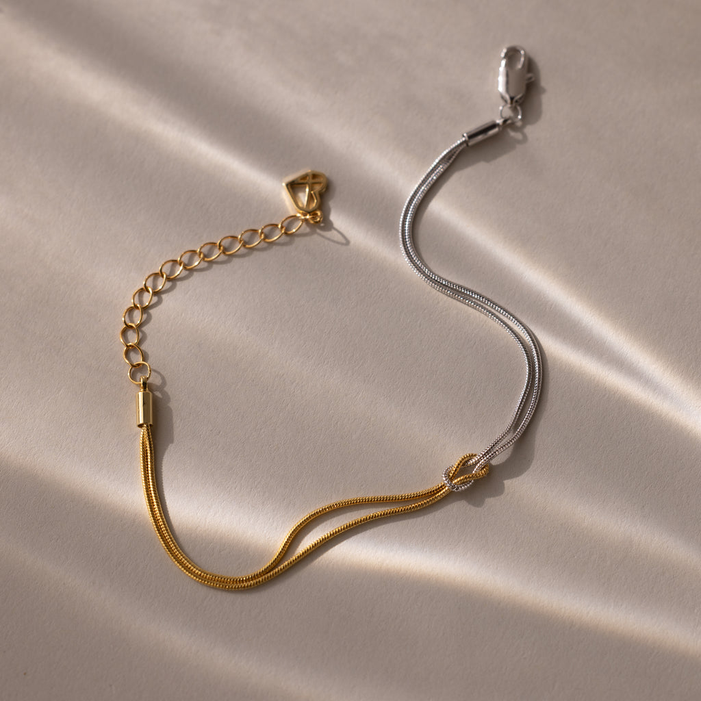 The Mixed Metal Knot Bracelet features gold and silver chains in a two-tone minimalist design, styled as a knot and displayed on a light, textured surface with soft shadows.