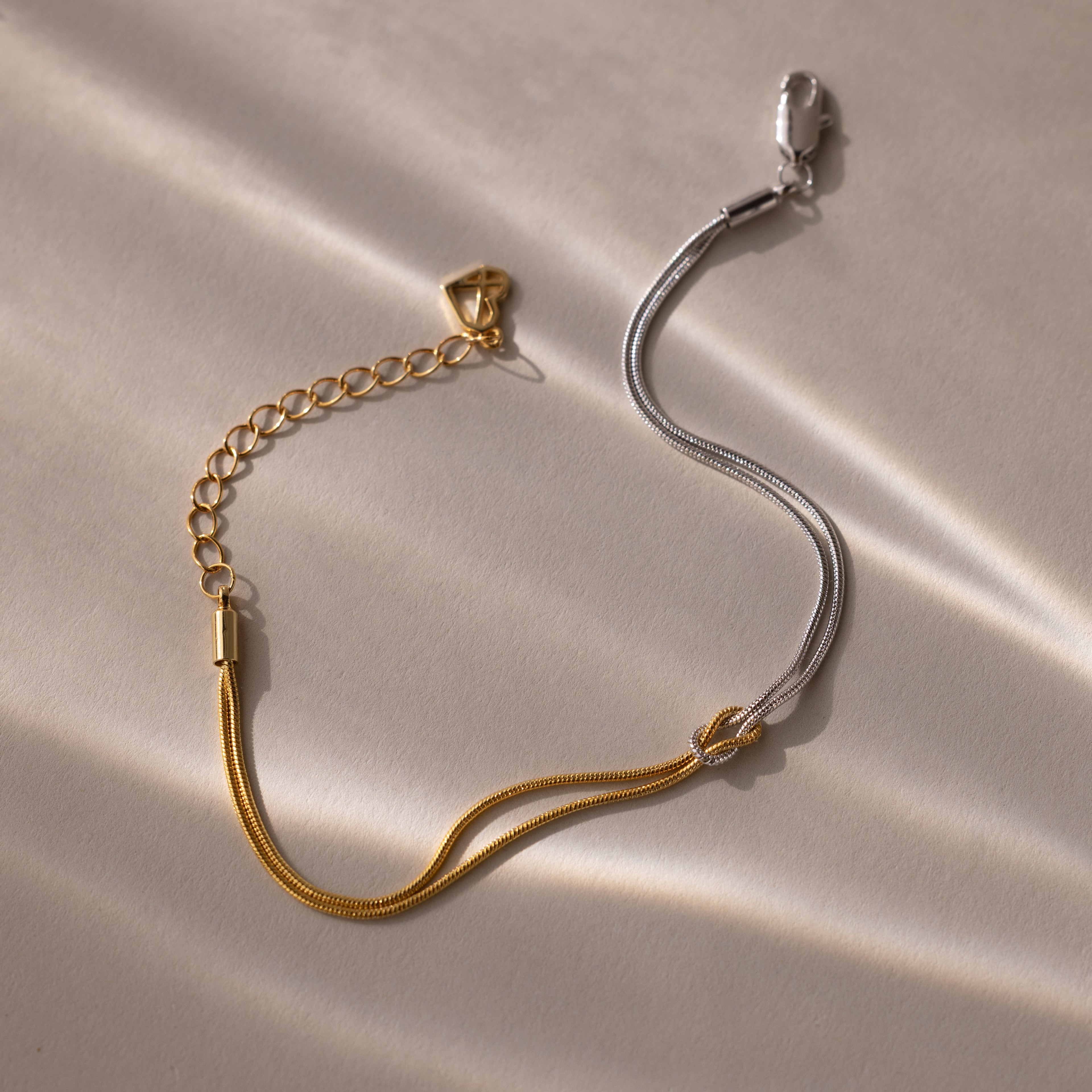 The Mixed Metal Knot Bracelet features gold and silver chains in a two-tone minimalist design, styled as a knot and displayed on a light, textured surface with soft shadows.