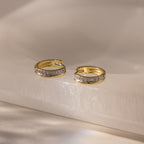 Half Pave Huggies in 18K Gold, featuring a row of sparkling CZ diamonds, are displayed on a smooth, light-colored surface.