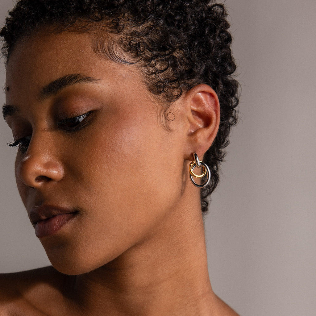 A woman with short curly hair wears Mixed Metal Linked Earrings in gold and silver, looking down against a neutral background.