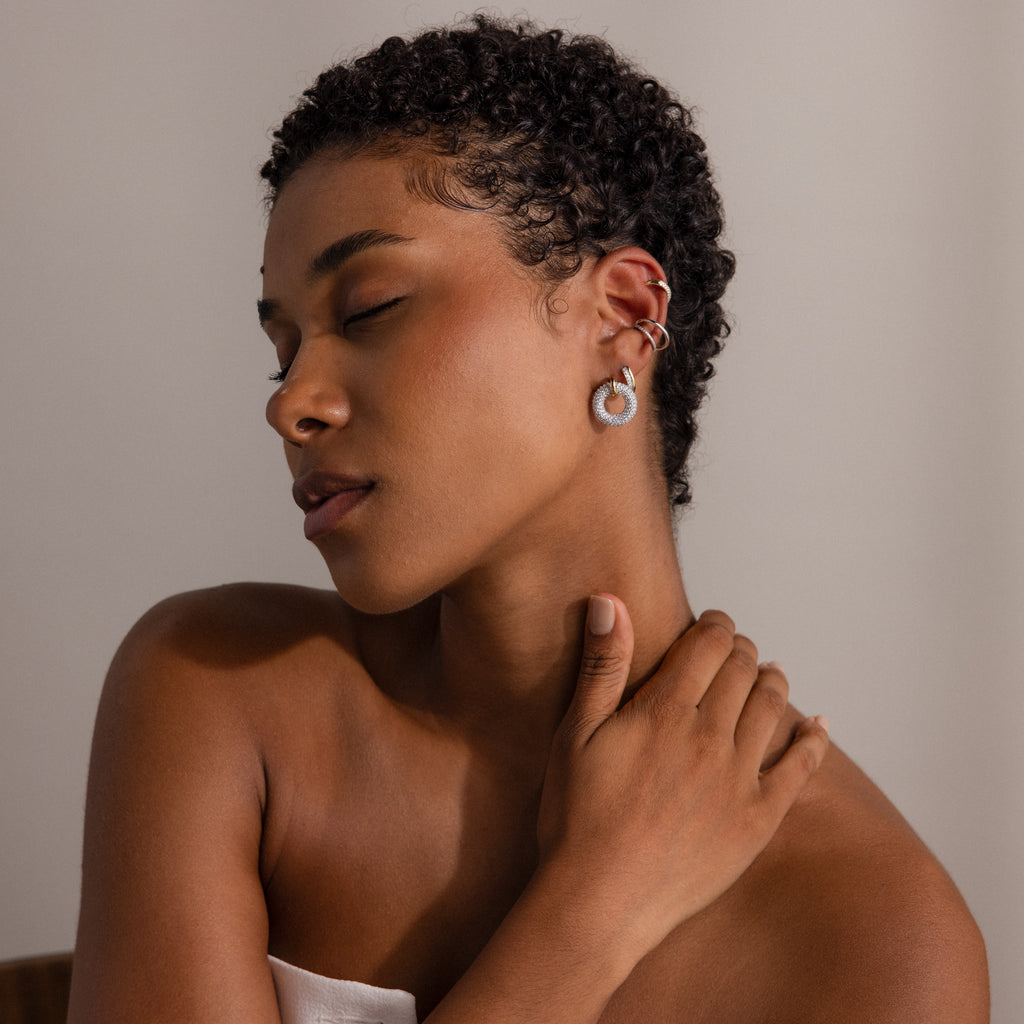 A woman with short curly hair, eyes closed and hand on neck, wears Half Pave Huggies earrings in gold and silver with CZ diamonds, her bare shoulders visible.