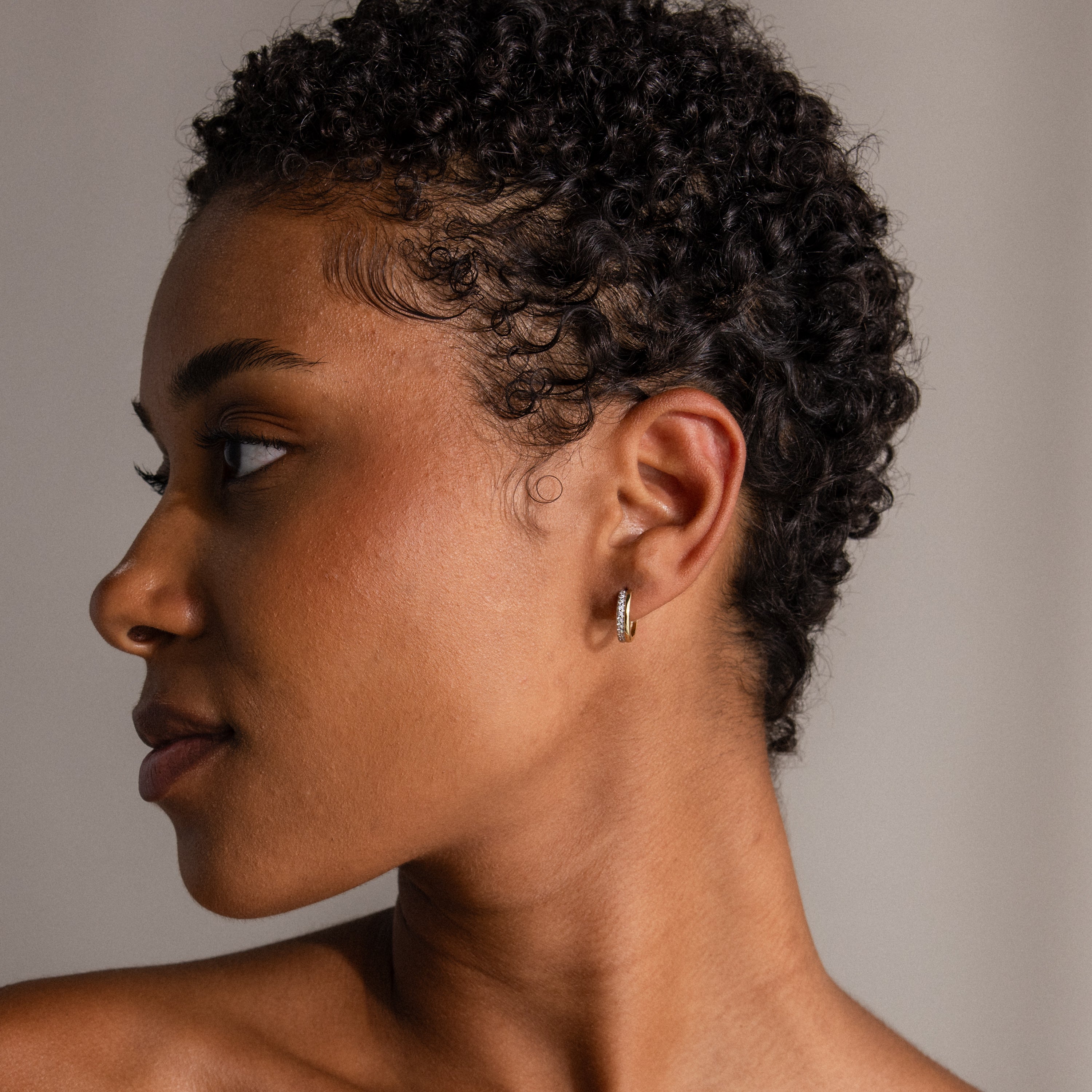 A woman with short curly hair looks left, displaying the Half Pave Huggies—small gold and silver earrings with CZ diamonds—against a neutral background.