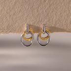 The Mixed Metal Linked Earrings feature interlocking gold and silver circles in a modern design, displayed on a textured beige background—perfect mixed metal hoops for a contemporary look.
