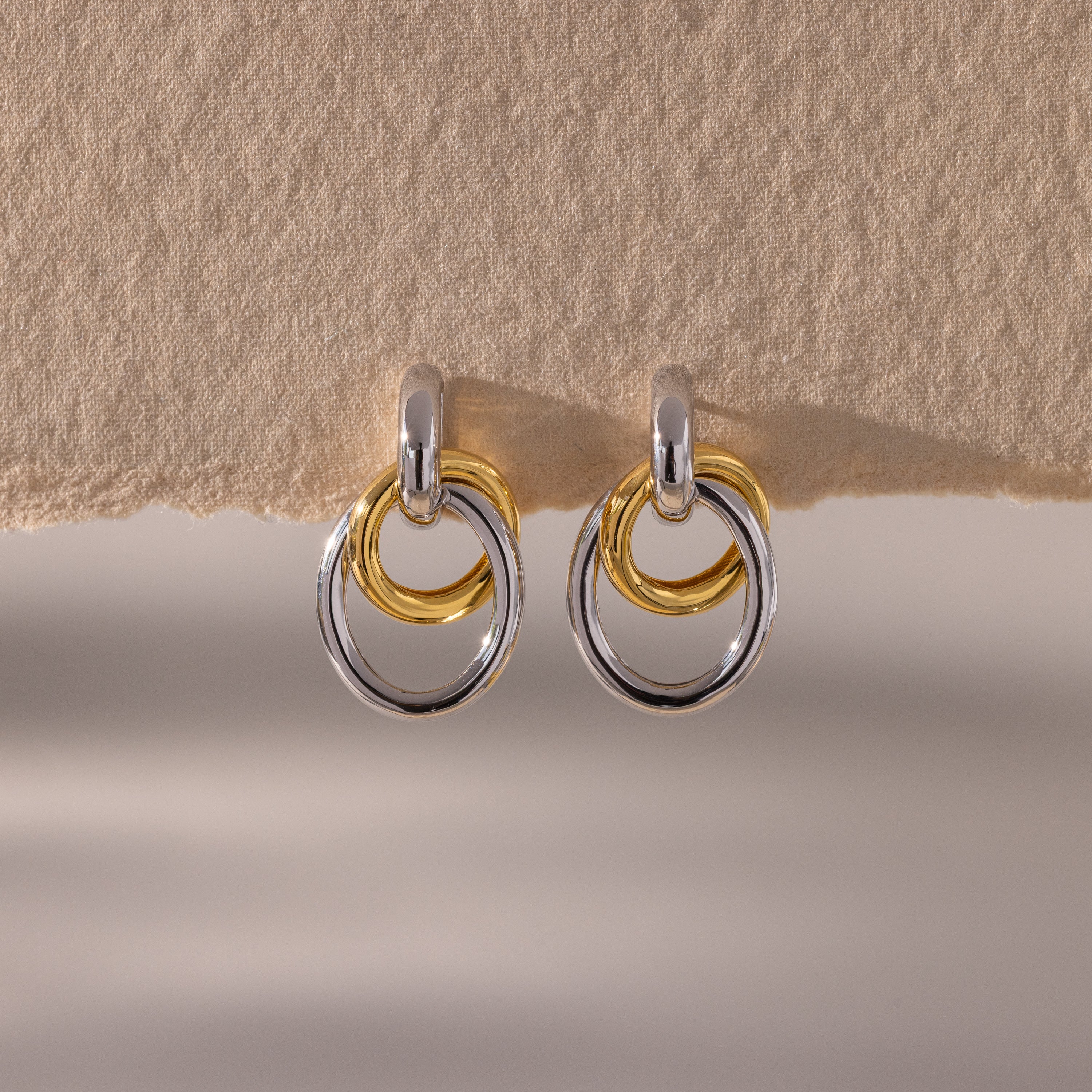 The Mixed Metal Linked Earrings feature interlocking gold and silver circles in a modern design, displayed on a textured beige background—perfect mixed metal hoops for a contemporary look.