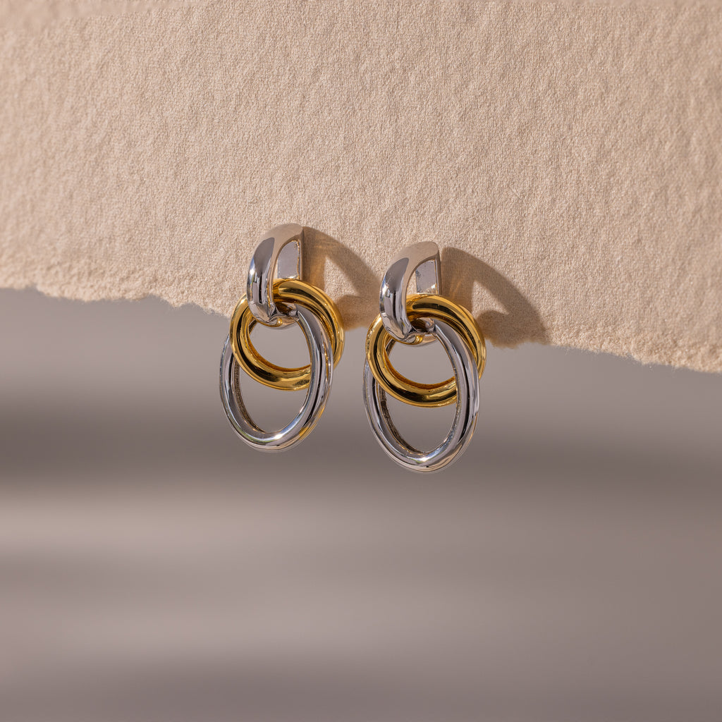 The Mixed Metal Linked Earrings feature gold and silver interlocking hoops in a modern design, showcased on a textured beige surface.
