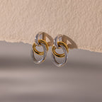 The Mixed Metal Linked Earrings feature gold and silver interlocking hoops in a modern design, showcased on a textured beige surface.