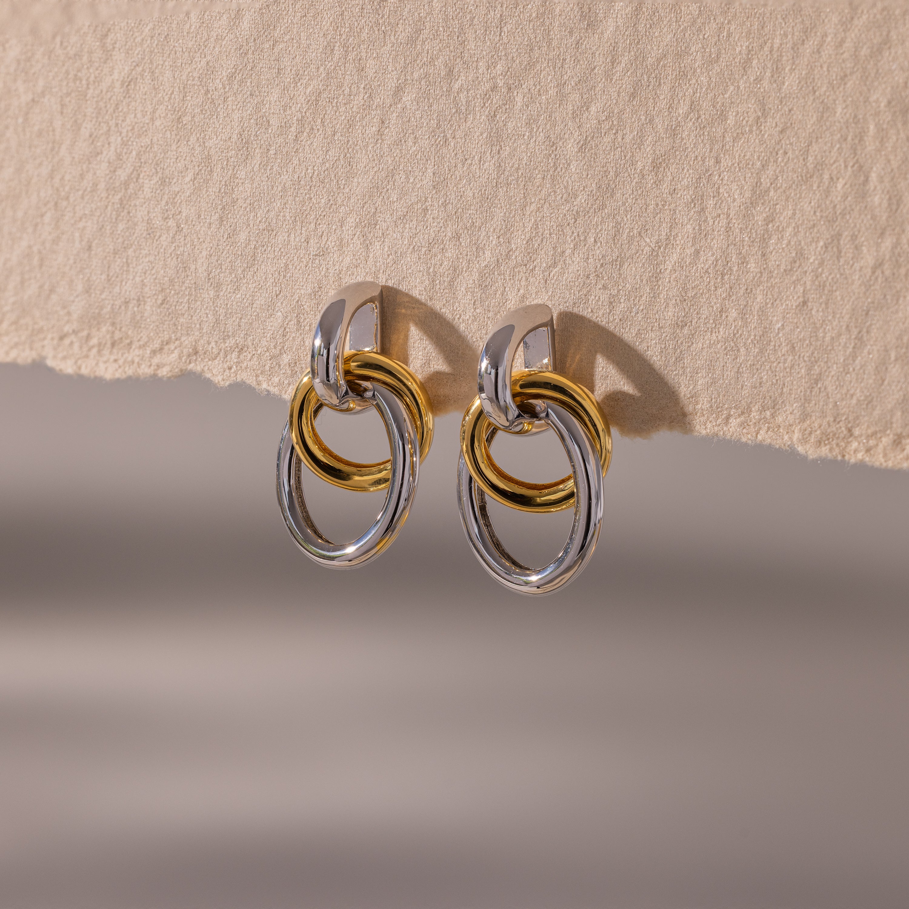 The Mixed Metal Linked Earrings feature gold and silver interlocking hoops in a modern design, showcased on a textured beige surface.