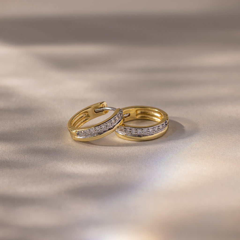 A pair of Half Pave Huggies in 18K Gold, featuring sparkling CZ stones, rests on a soft, neutral surface under gentle lighting.