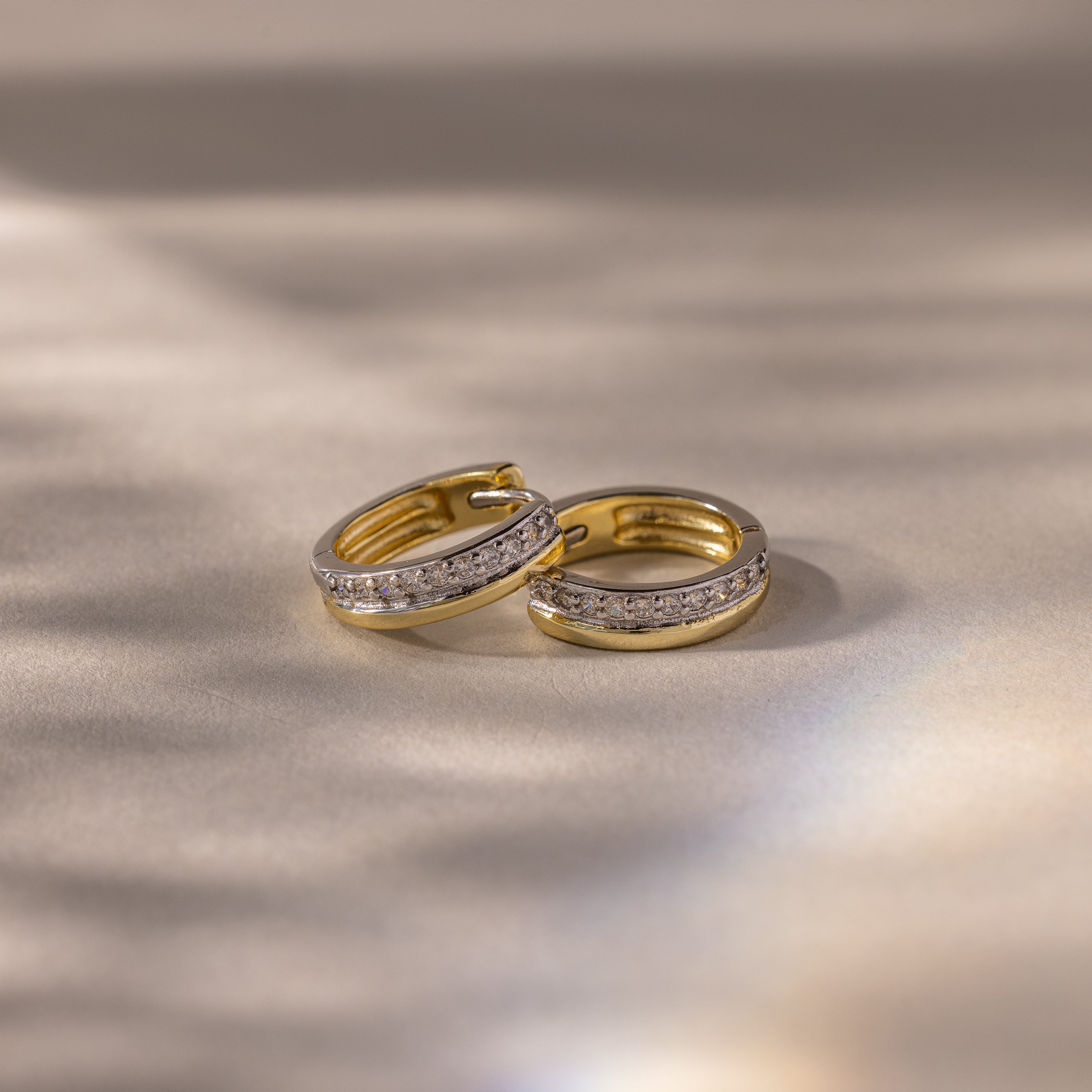 Half Pave Huggies in Mixed Metal hoop earrings with embedded CZ diamonds, displayed on a soft, shimmering beige surface.