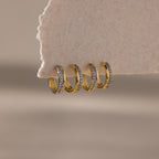 Four Half Pave Huggies gold hoop earrings, each adorned with small CZ diamonds, hang on textured beige paper, showcasing their modern design.