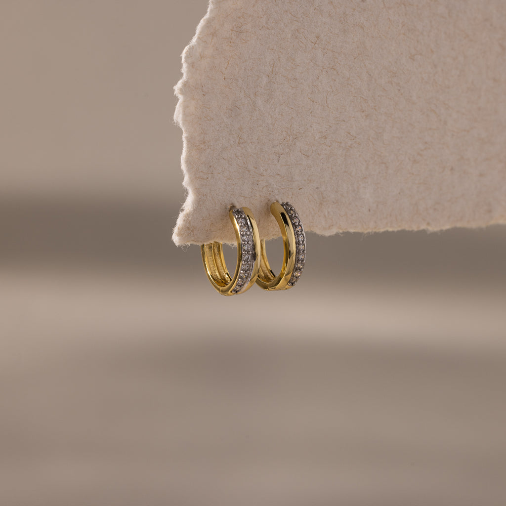 The Half Pave Huggies in 18K Gold, adorned with sparkling CZ stones, hang elegantly on a textured beige paper edge.
