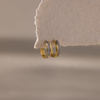 The Half Pave Huggies in 18K Gold, adorned with sparkling CZ stones, hang elegantly on a textured beige paper edge.