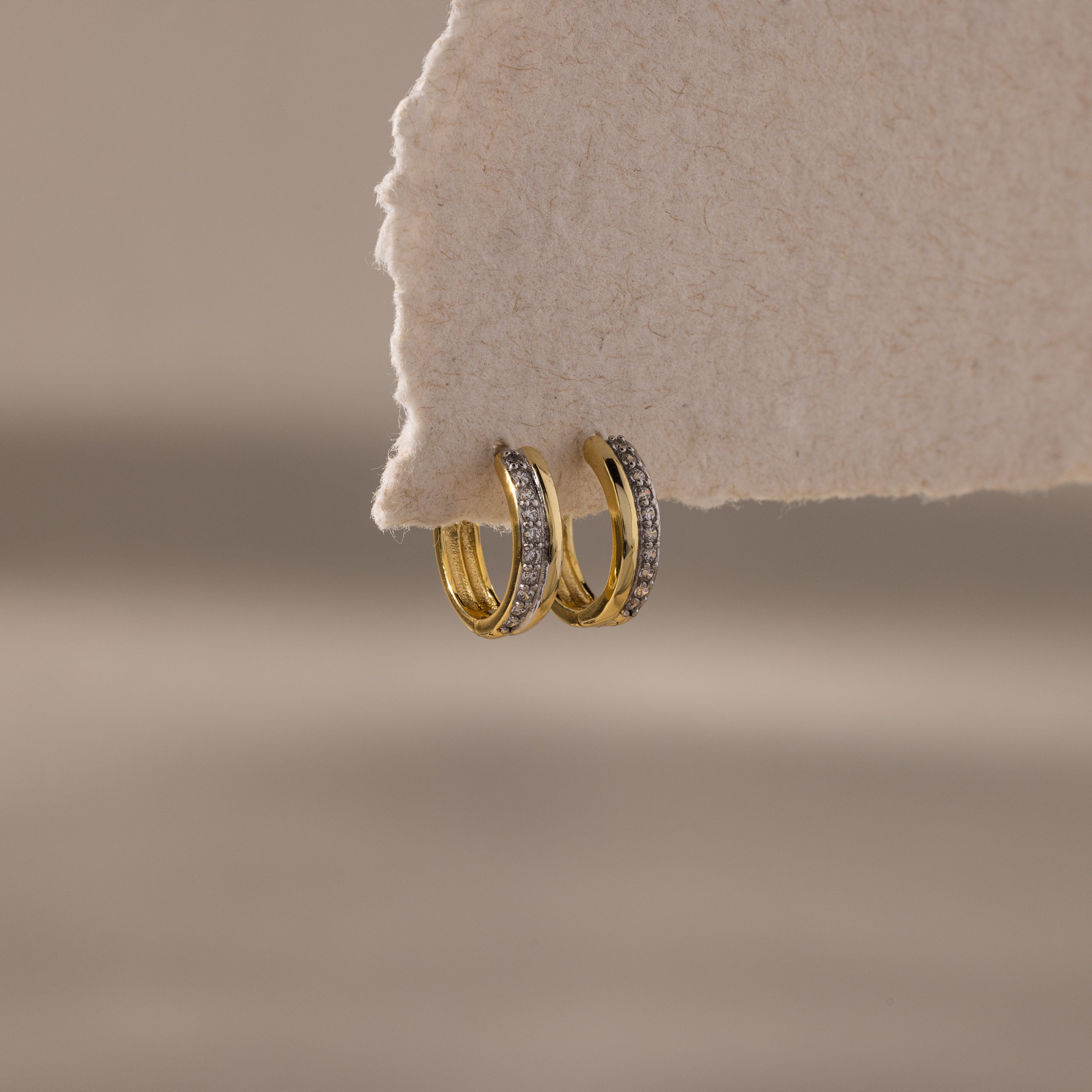 The Half Pave Huggies in 18K Gold, adorned with sparkling CZ stones, hang elegantly on a textured beige paper edge.