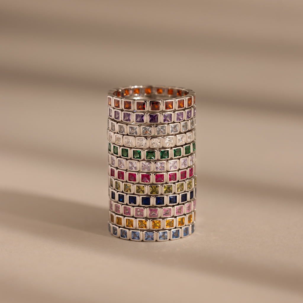 A stack of Eternity Bezel Birthstone Rings with square-cut gemstones in elegant bezel settings, arranged in rows on a beige surface with soft shadow lines.