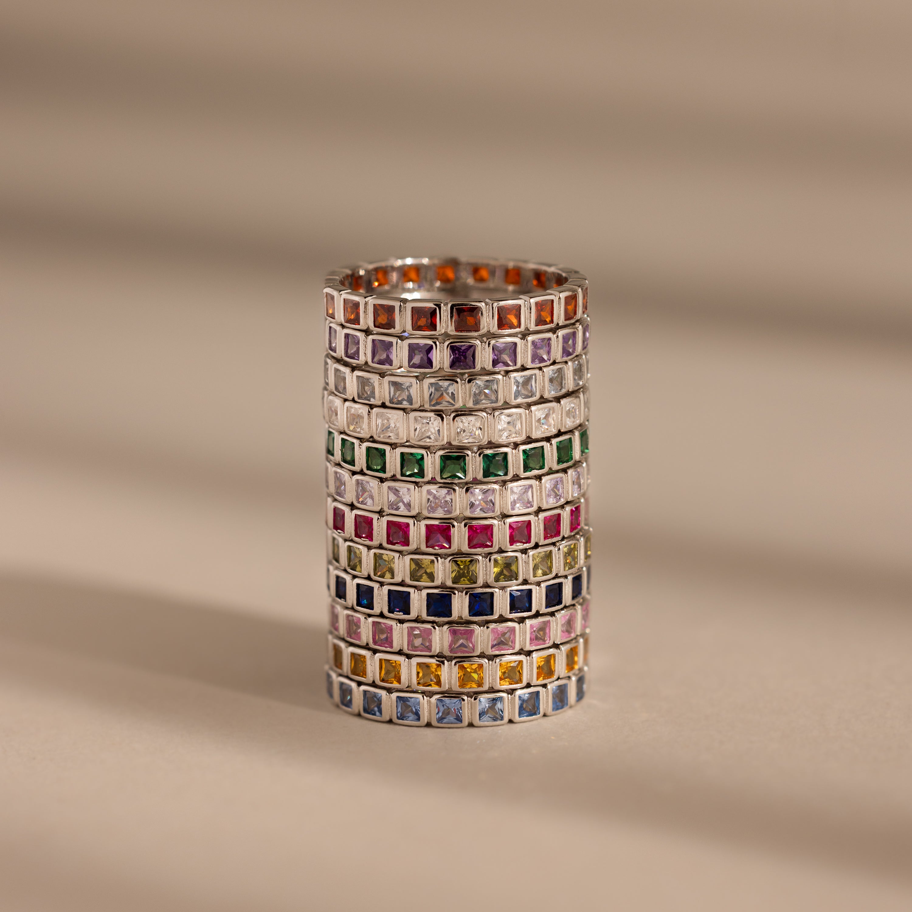 A stack of Eternity Bezel Birthstone Rings with square-cut gemstones in elegant bezel settings, arranged in rows on a beige surface with soft shadow lines.