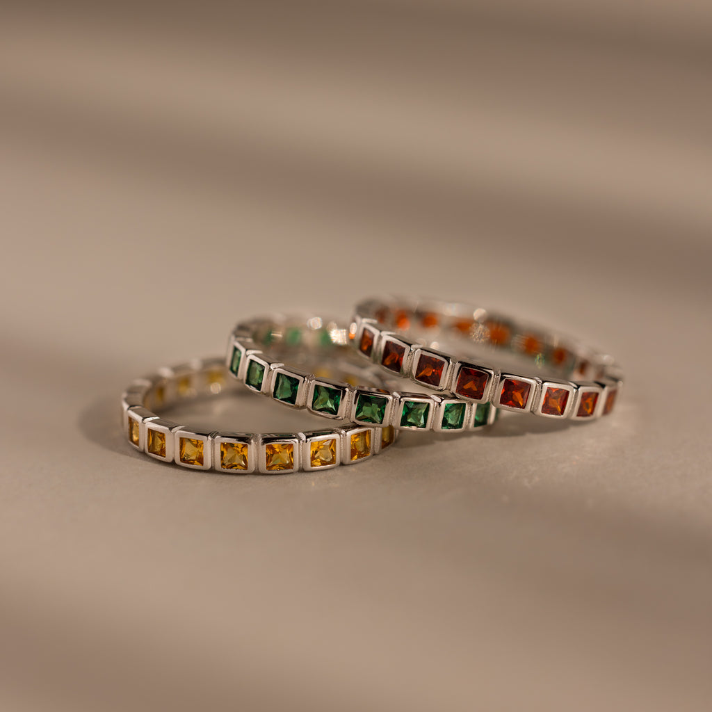 Three Eternity Bezel Birthstone Rings, each with a square cut gemstone in yellow, green, and orange bezel settings, are stacked elegantly on a neutral surface.