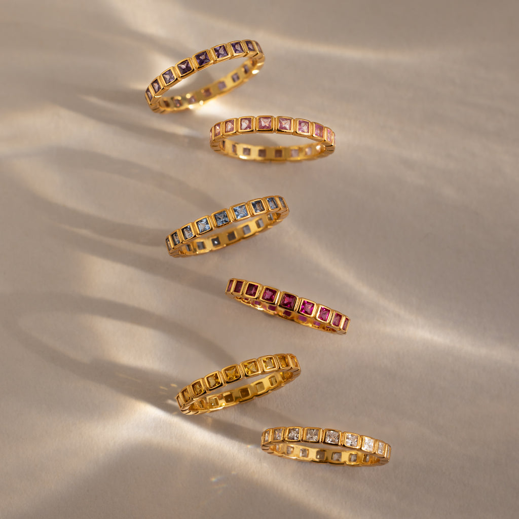 Six gold Eternity Bezel Garnet Rings with square cut garnet gemstones are arranged diagonally on a neutral background.