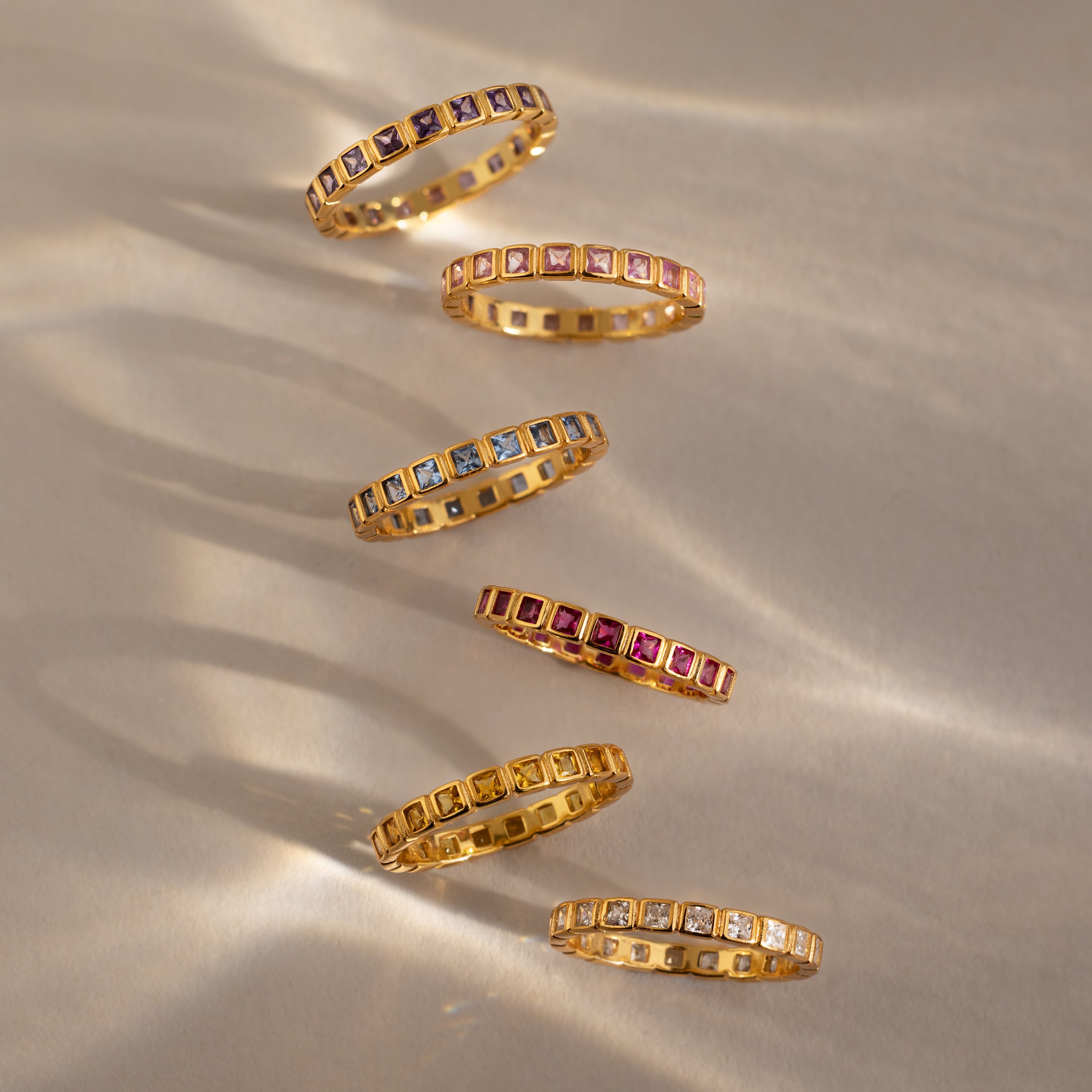 Six gold Eternity Bezel Garnet Rings with square cut garnet gemstones are arranged diagonally on a neutral background.