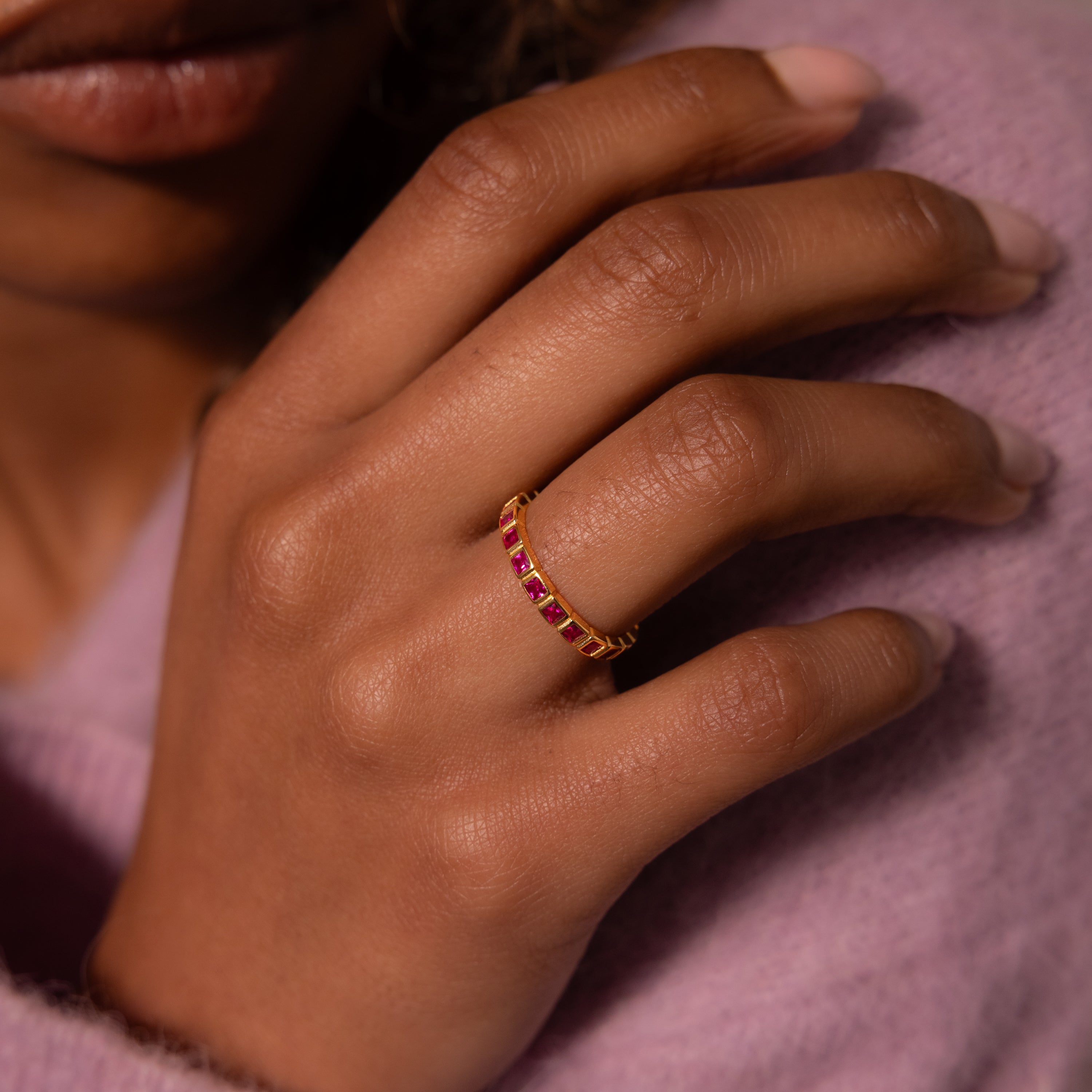 A hand wearing the Eternity Bezel Birthstone Ring, featuring square-cut red gemstones in a bezel setting, rests on a lavender sweater.