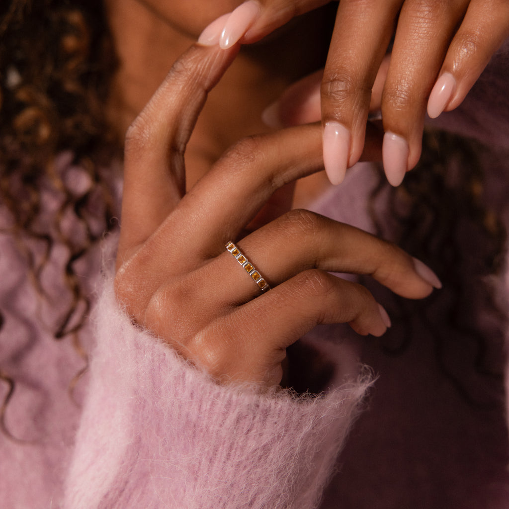 A hand with pale pink nails wearing a gold Eternity Bezel Garnet Ring, shown against a soft pink fuzzy sweater.