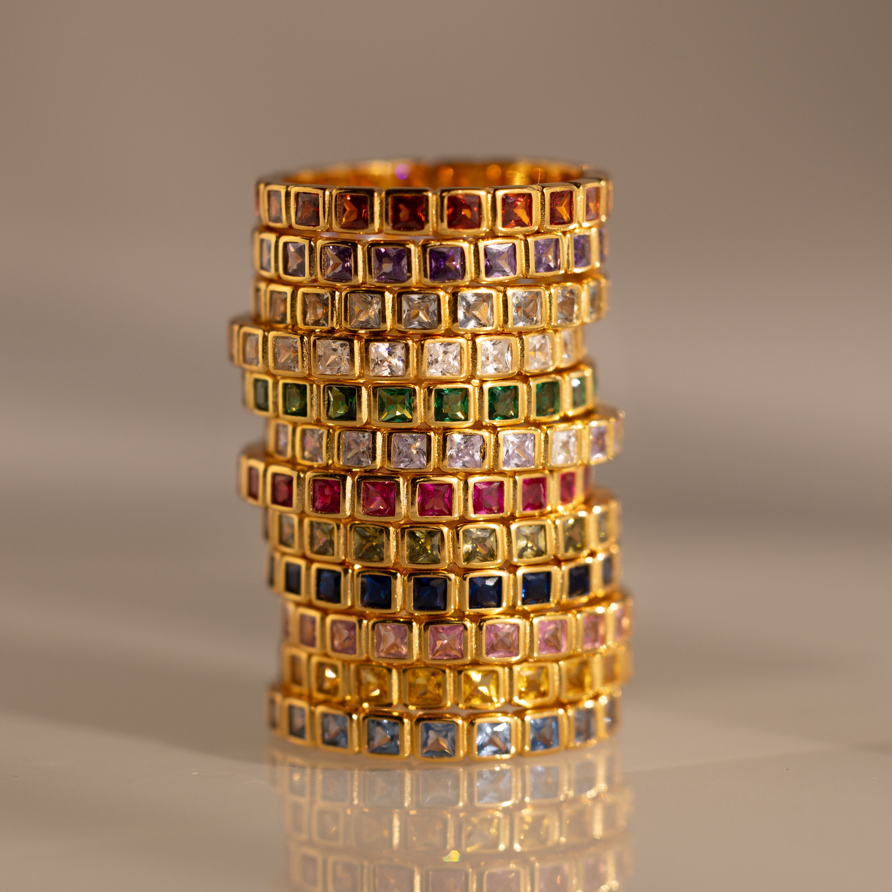 A stack of Eternity Bezel Birthstone Rings with square-cut gemstones in bezel settings is arranged in rows on a reflective surface.
