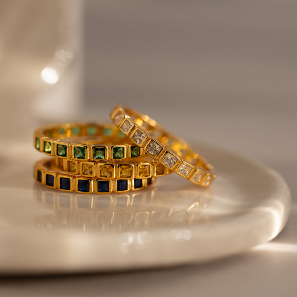 The Eternity Bezel Emerald Ring, a gold ring with a green square-cut gemstone in a bezel setting, is elegantly stacked alongside other gemstone rings on a white ceramic dish.