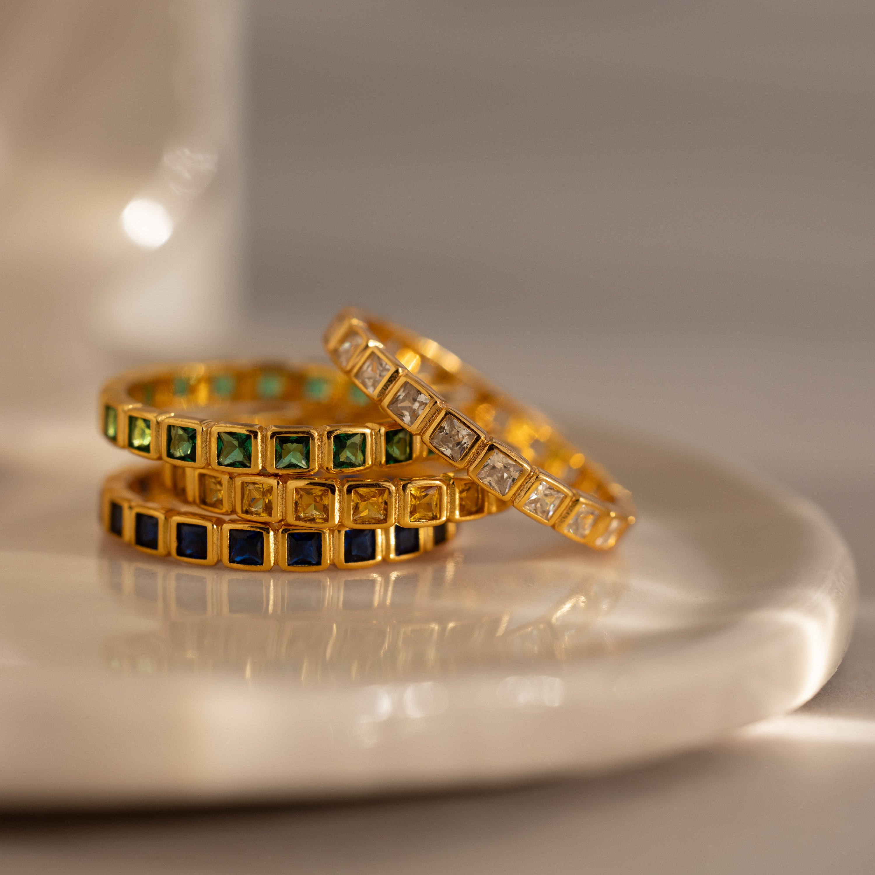 The Eternity Bezel Emerald Ring, a gold ring with a green square-cut gemstone in a bezel setting, is elegantly stacked alongside other gemstone rings on a white ceramic dish.