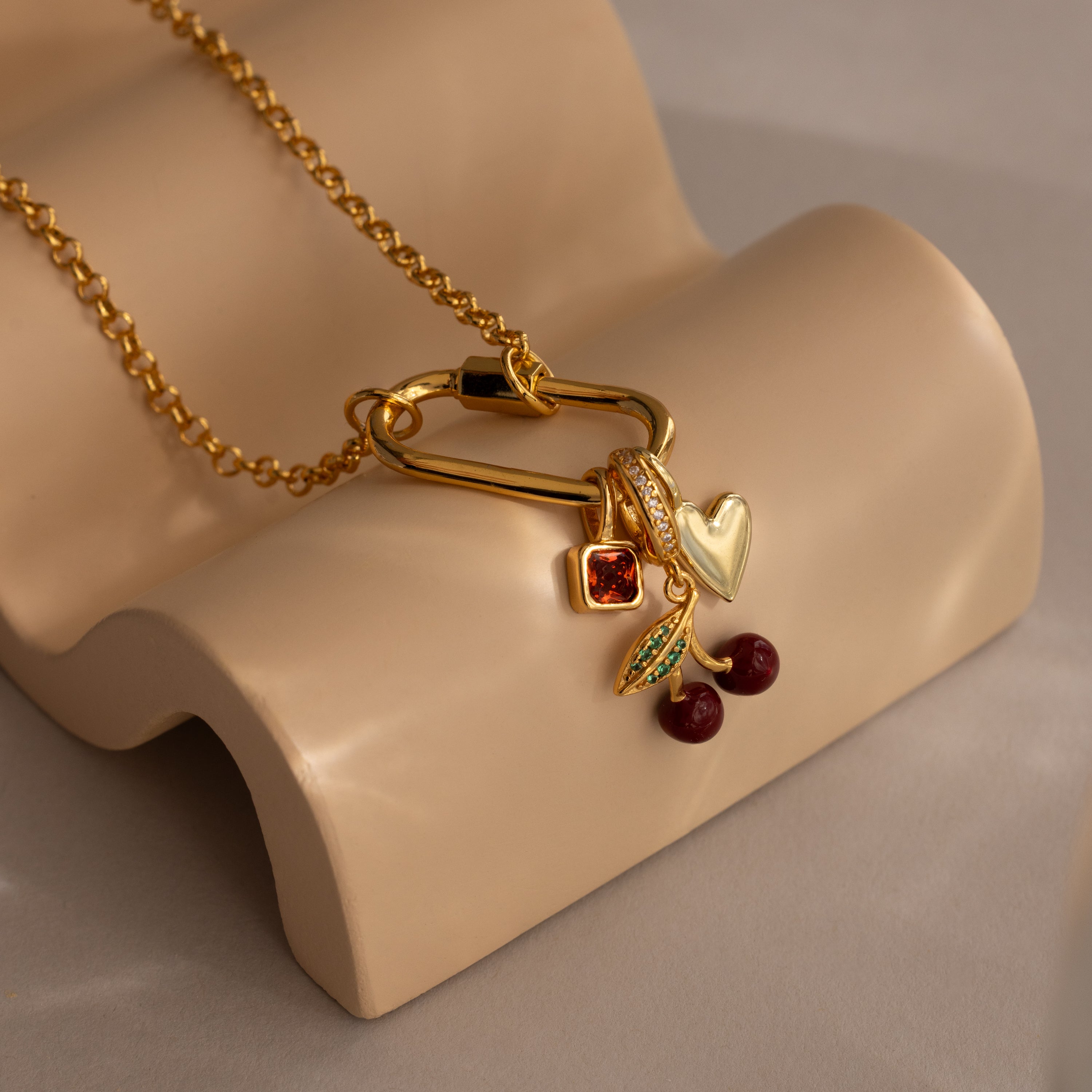 Our Ruby Cherry Charm from the jewelry collection showcases a heart, cherries, a red pavé stone, and a leafy branch on beige—adding vibrant elegance to any look.