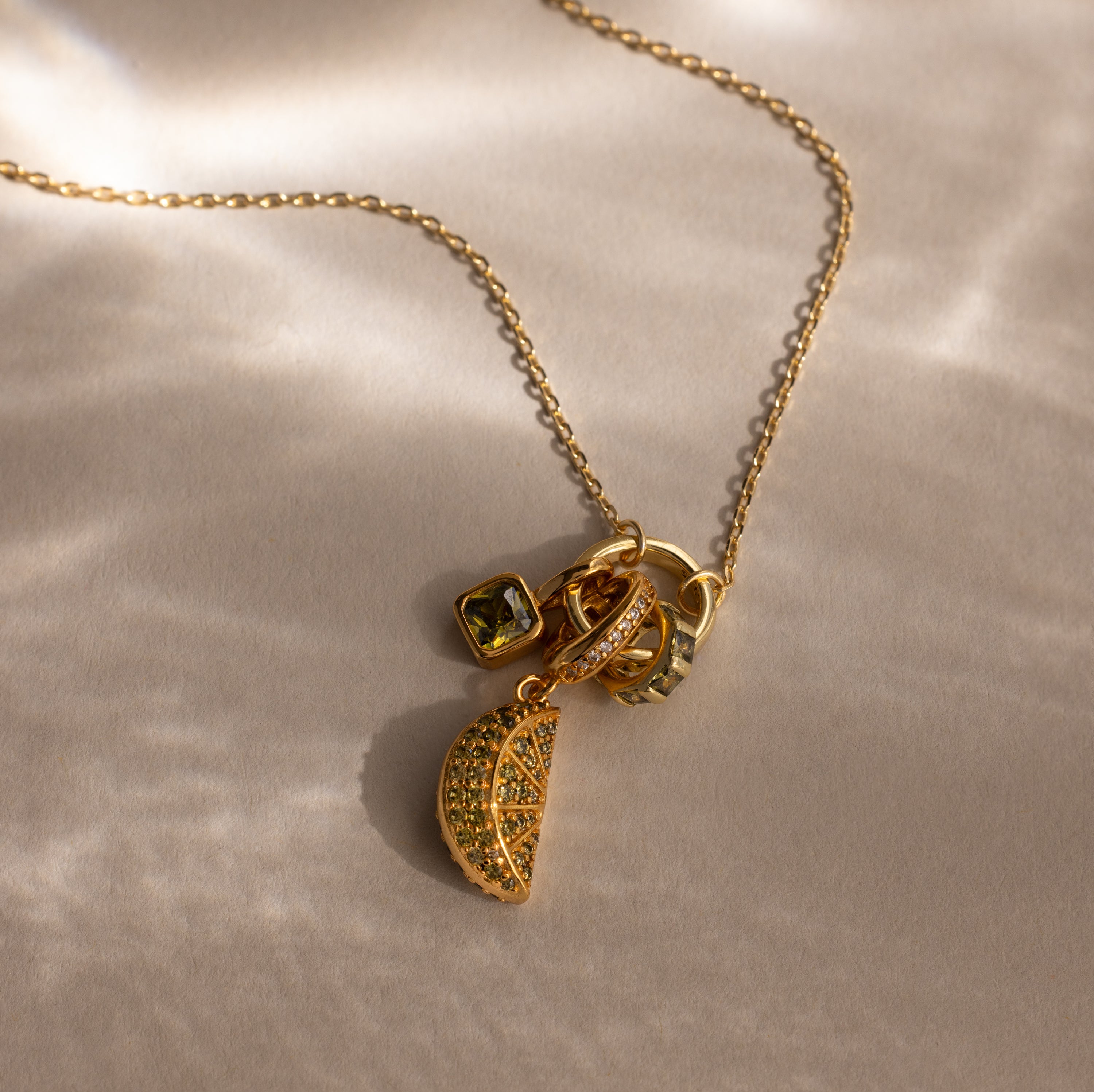 The Birthstone Fruit Charm, a gold necklace featuring a jeweled lemon slice pavé stone charm, is displayed on a soft beige surface in sunlight—a standout piece from our jewelry collection.