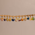 The Birthstone Fruit Charm features gold, fruit-shaped charms—pavé stone strawberries, bananas, and cherries—dangling from a wooden branch on a beige background, adding playful flair to any jewelry collection.