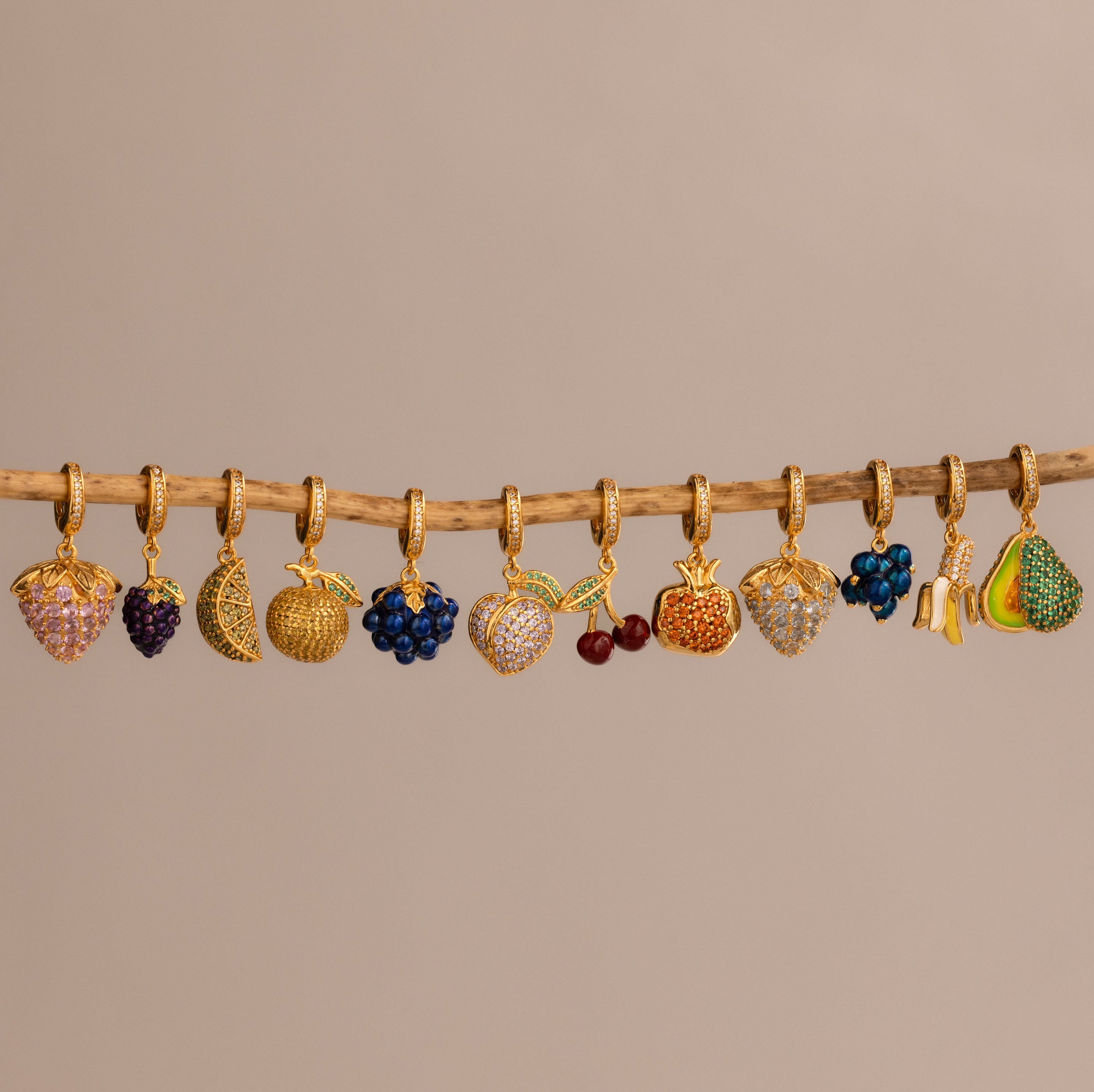 The Birthstone Fruit Charm features gold, fruit-shaped charms—pavé stone strawberries, bananas, and cherries—dangling from a wooden branch on a beige background, adding playful flair to any jewelry collection.